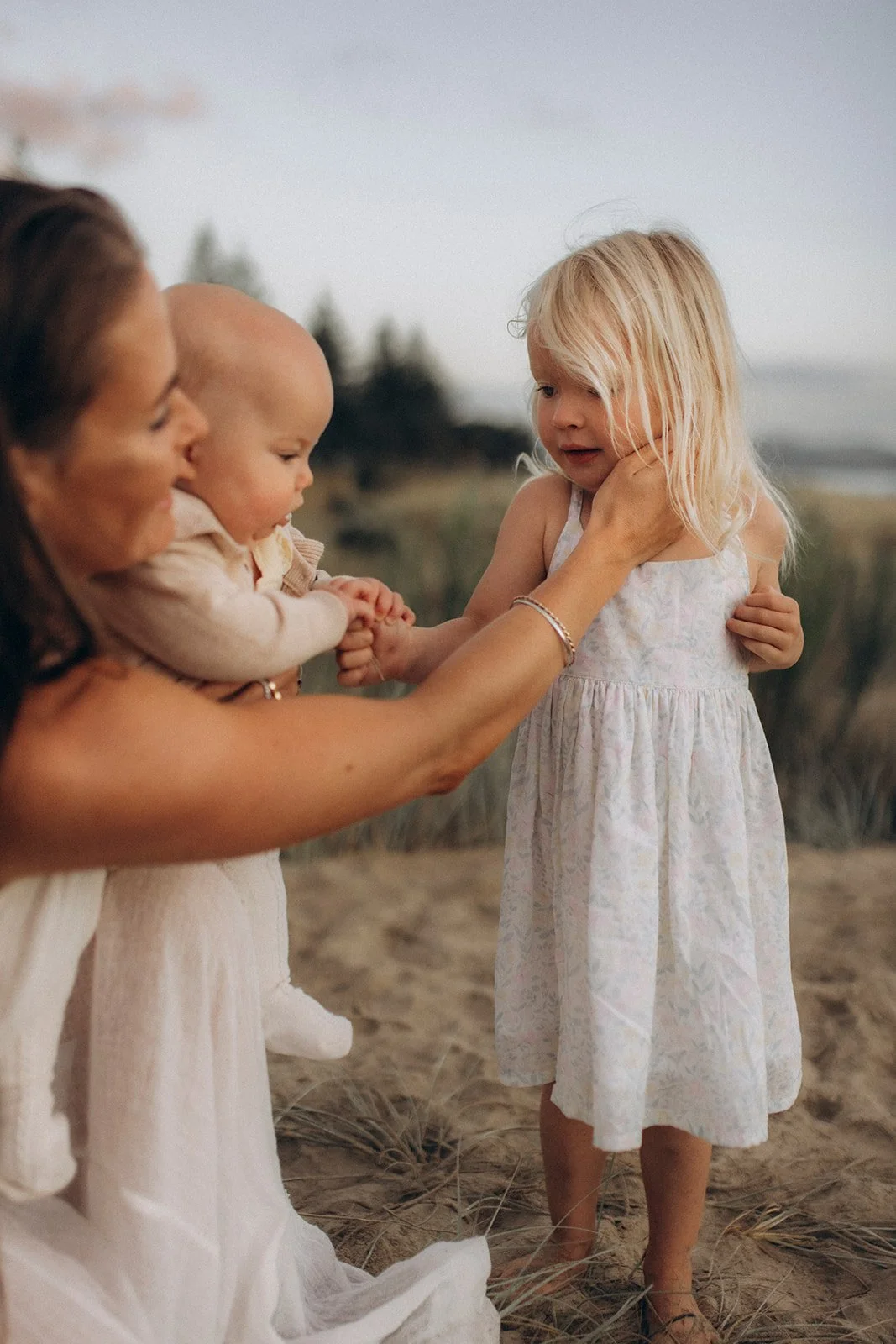 A woman with brown hair and a white dress is holding a smiling baby while touching the cheek of a little blonde girl in a white dress on a beach with sand and grass, behind them water and trees are visible.
