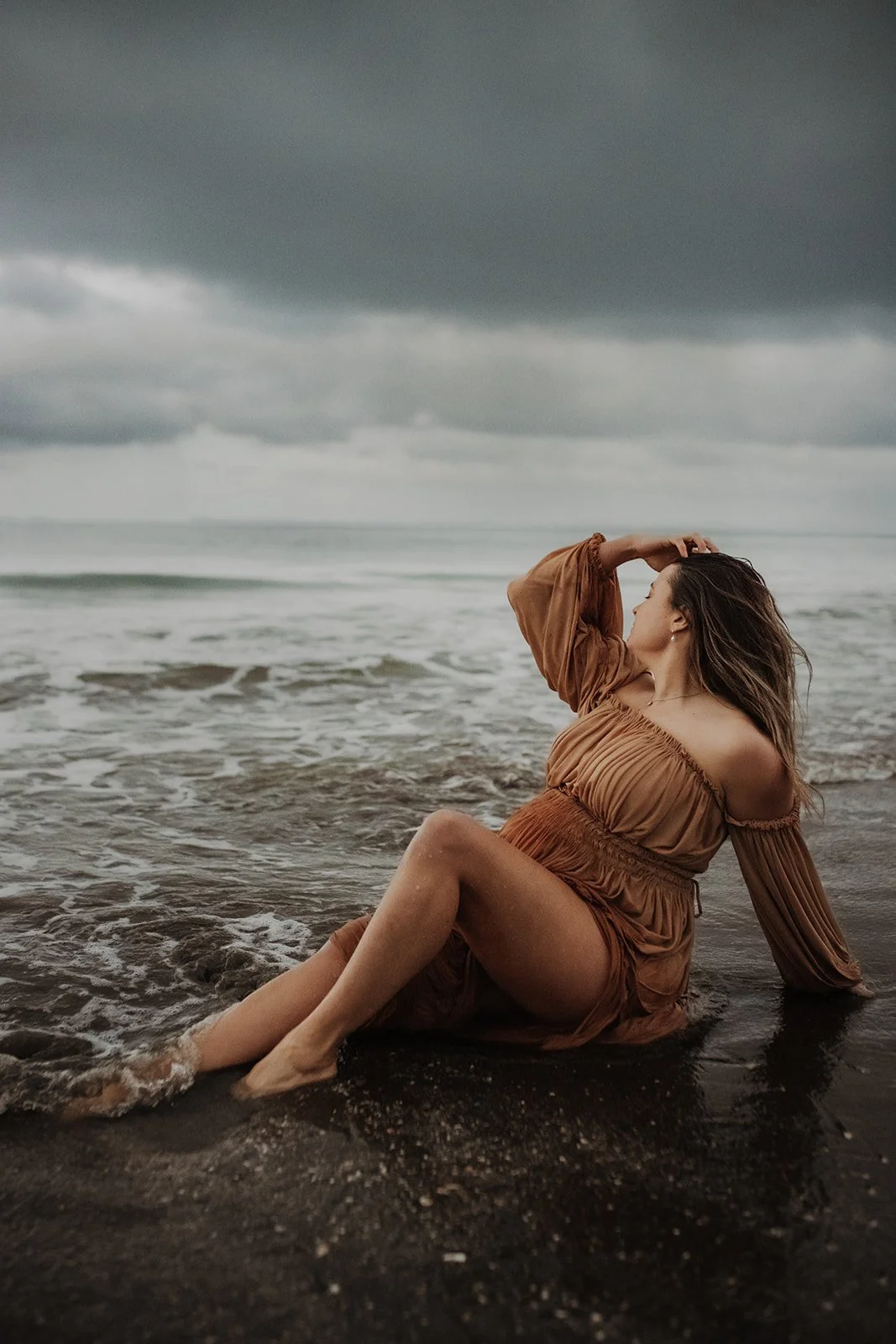 Woman in a brown dress sitting on a wet beach with waves around her under a cloudy sky.