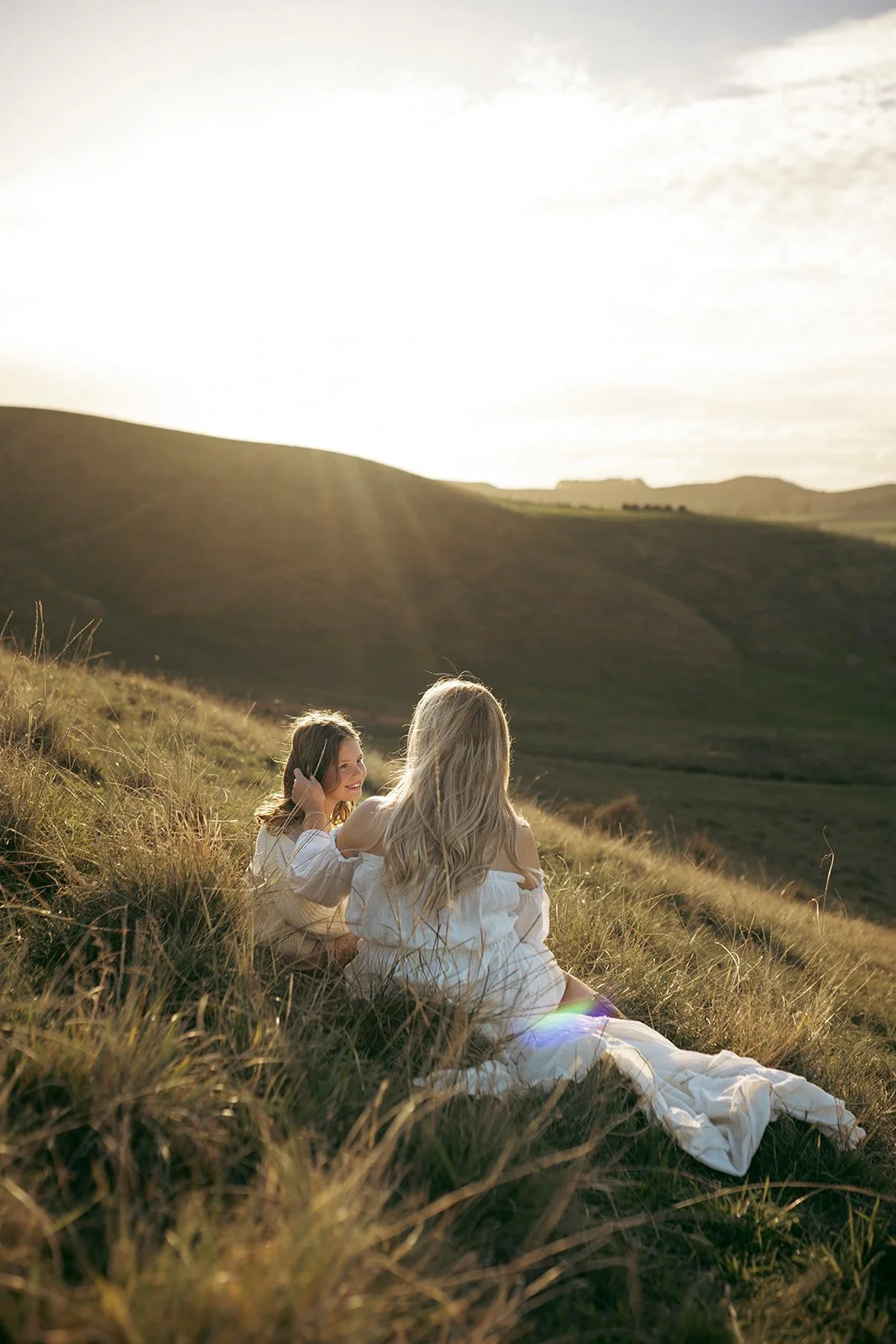 Two women sitting on a grassy hillside at sunset, wearing white dresses, with hills in the background.