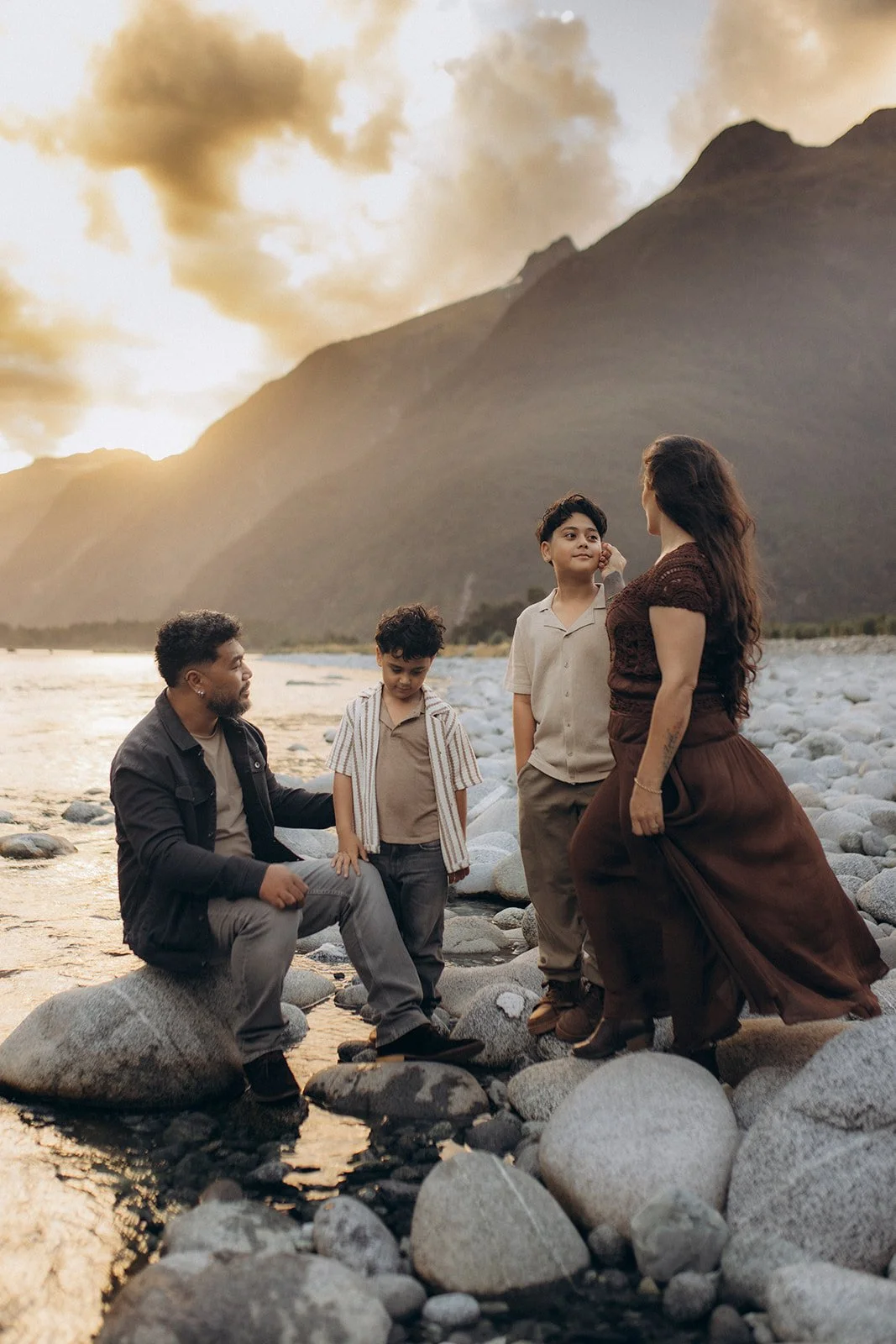 A family of four having a conversation on a rocky riverbank at sunset with mountains in the background.