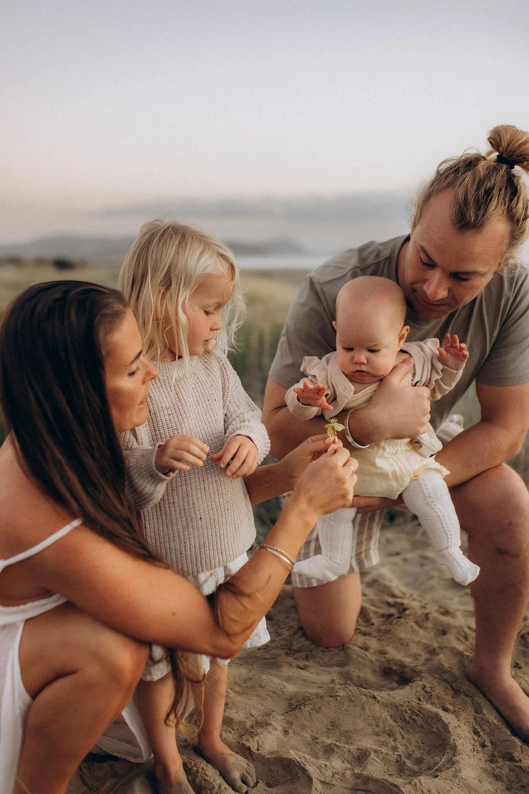 A family on the beach, with a woman, two young girls, and a man holding a baby, all focused on a small flower in the woman's hand.