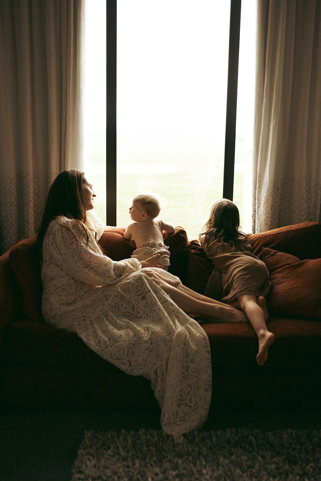 A woman and two children sitting on a couch in front of a large window with curtains. The woman is smiling at a young boy, and a girl is leaning over the back of the couch facing away from the camera.
