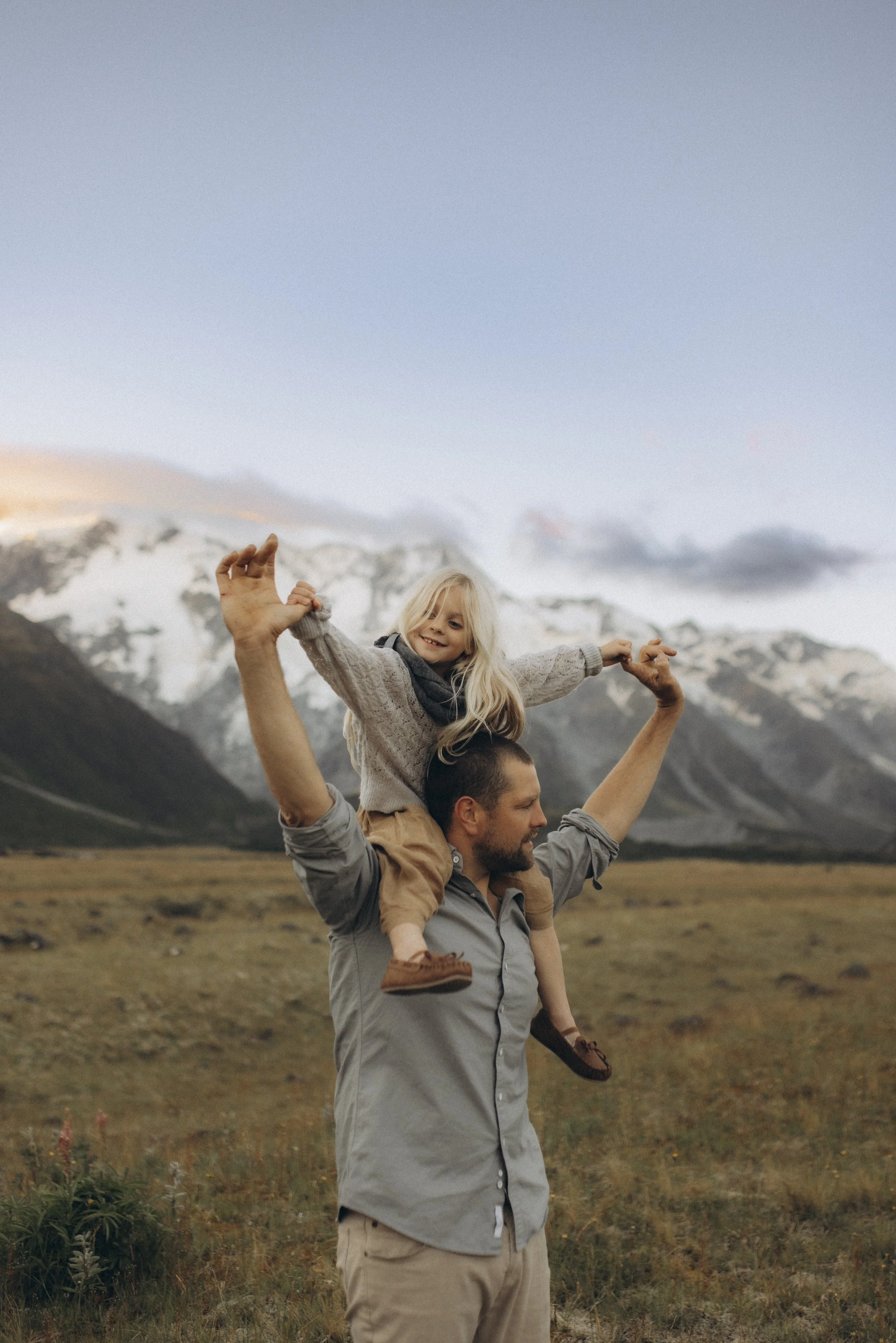 A man is holding a young girl on his shoulders in a mountain landscape with snow-capped peaks and a grassy field.