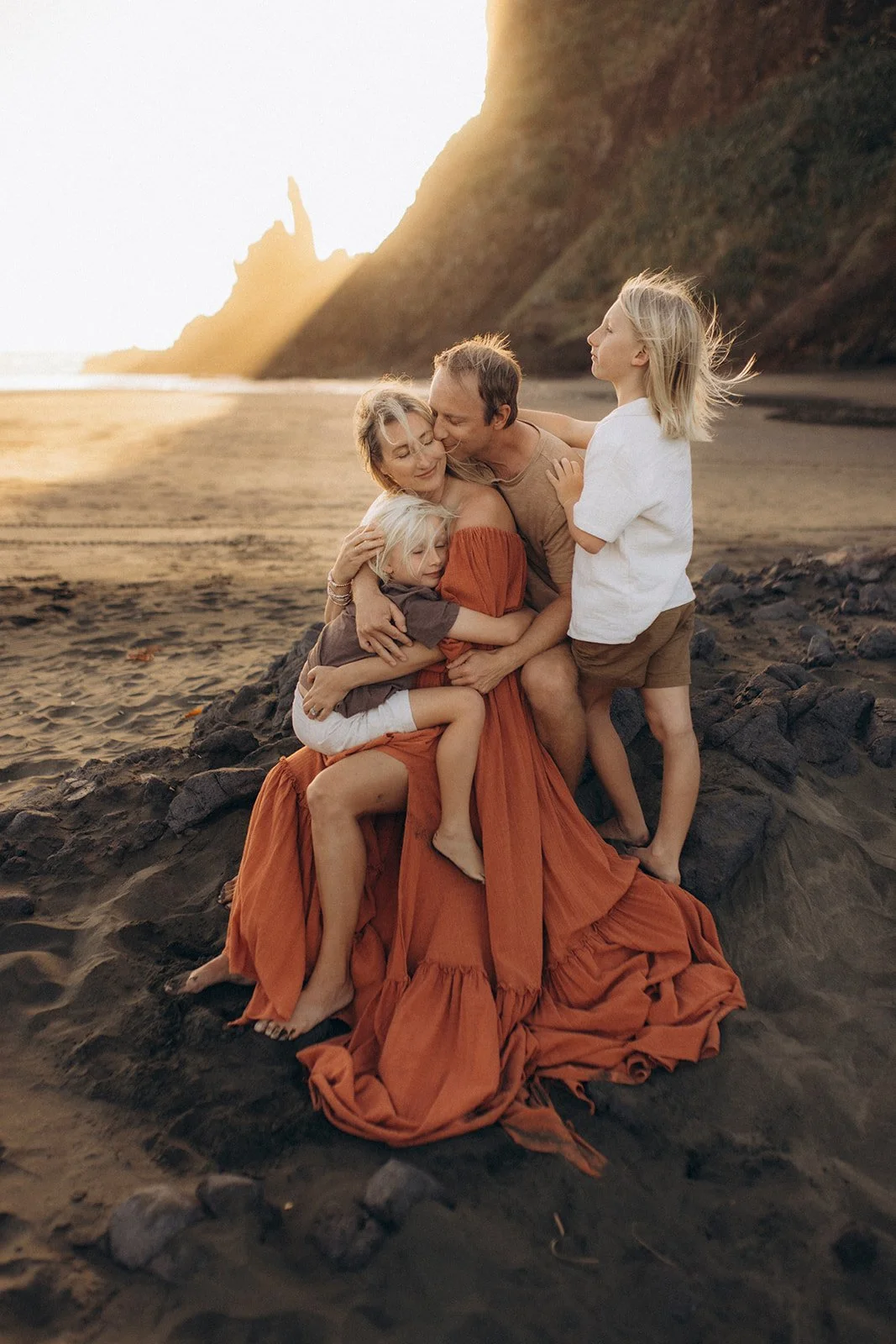 A family sitting on rocks at the beach during sunset, hugging and enjoying each other's company.