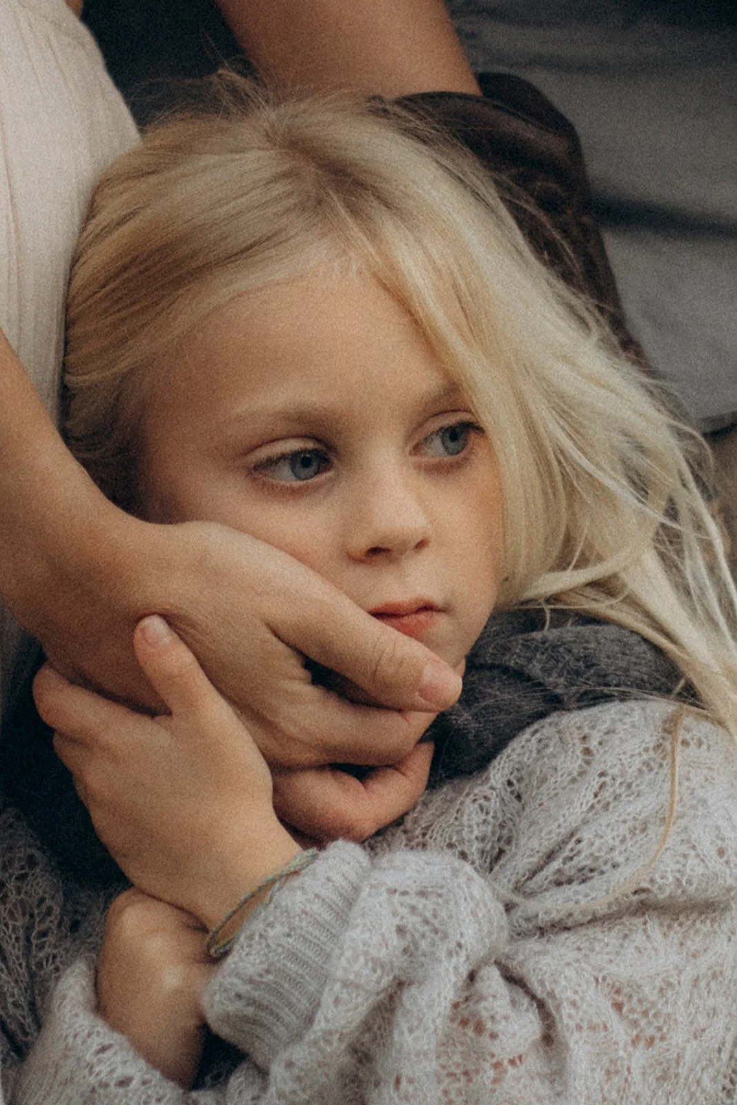 A young girl with blonde hair and blue eyes is being gently held by an adult's hand near her face.