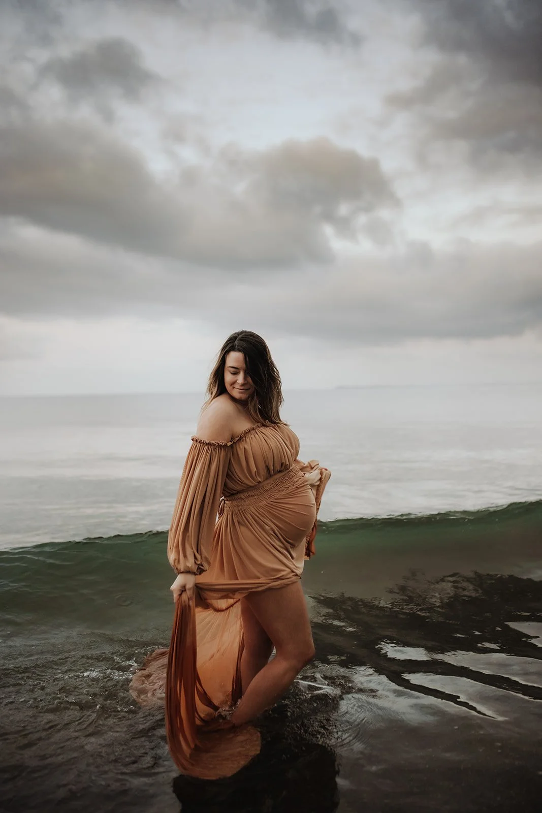 A pregnant woman standing in shallow ocean water, wearing a flowing beige dress, under a cloudy sky.