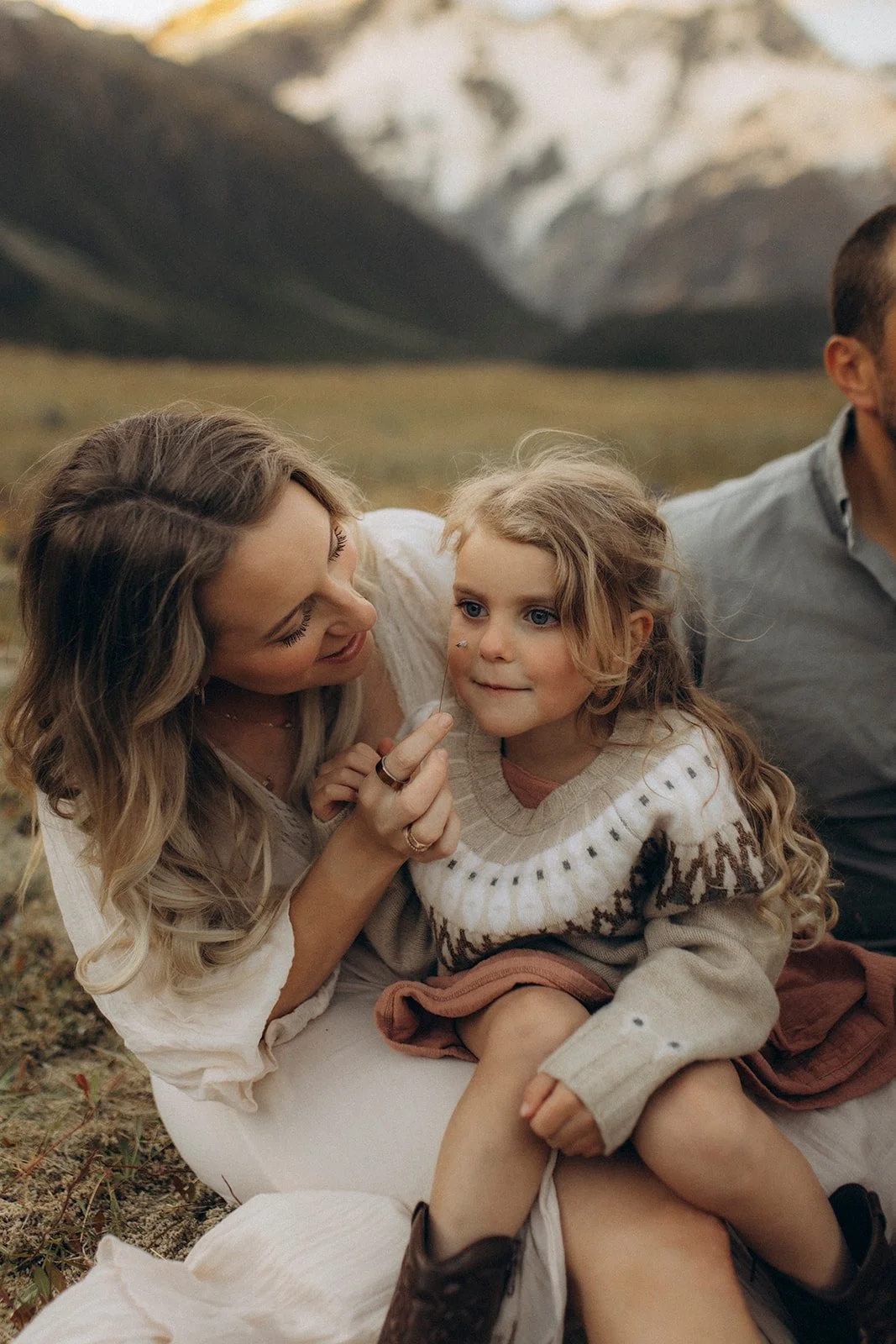 A woman and a girl sitting outdoors in a grassy area with mountains in the background. The woman is playfully using a stick or a flower on the girl's face, and both are smiling. The girl has long, curly blonde hair and is wearing a beige sweater with