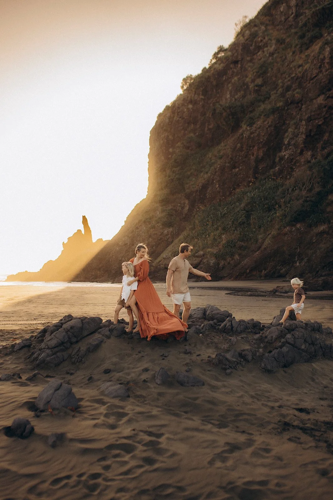 Family of four on a rocky beach during sunset, with a cliff in the background. The mother and children are on a small rocky outcrop, with the mother in an orange dress holding a young girl, and a man walking nearby with a young girl sitting on the ro