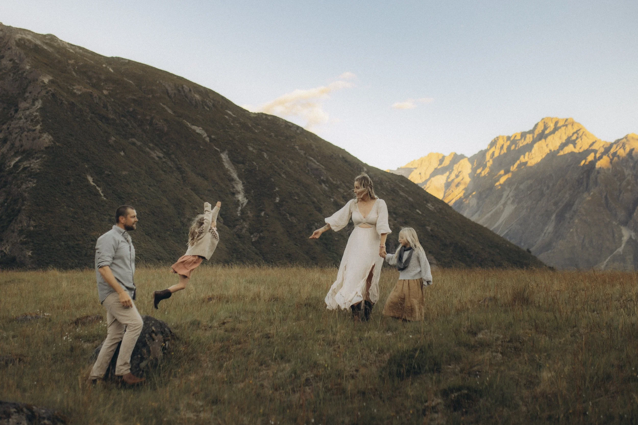 A family of five enjoying time together in a grassy field with mountains in the background during sunset, with children playing and a woman holding hands with a young girl.