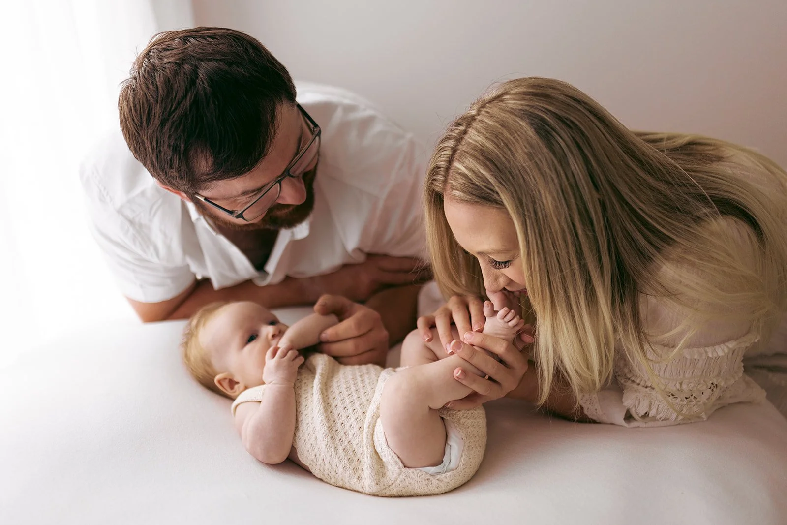 A family of three with a baby lying on a bed: a woman, a man, and a baby. The woman and the baby are holding hands, and the woman is kissing the baby's foot.