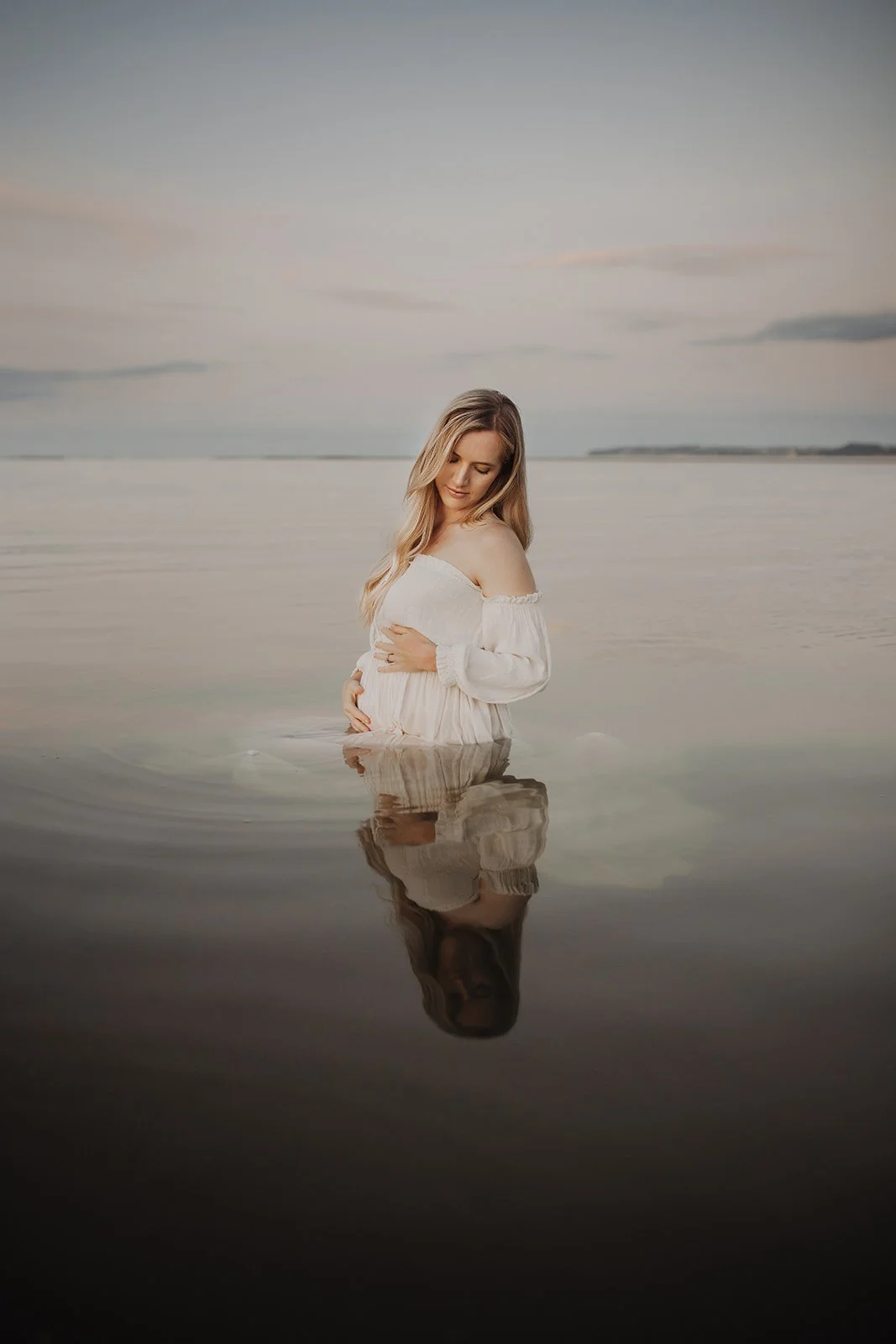 A pregnant woman in a white dress standing in shallow water at the beach during twilight, with her reflection visible in the water.