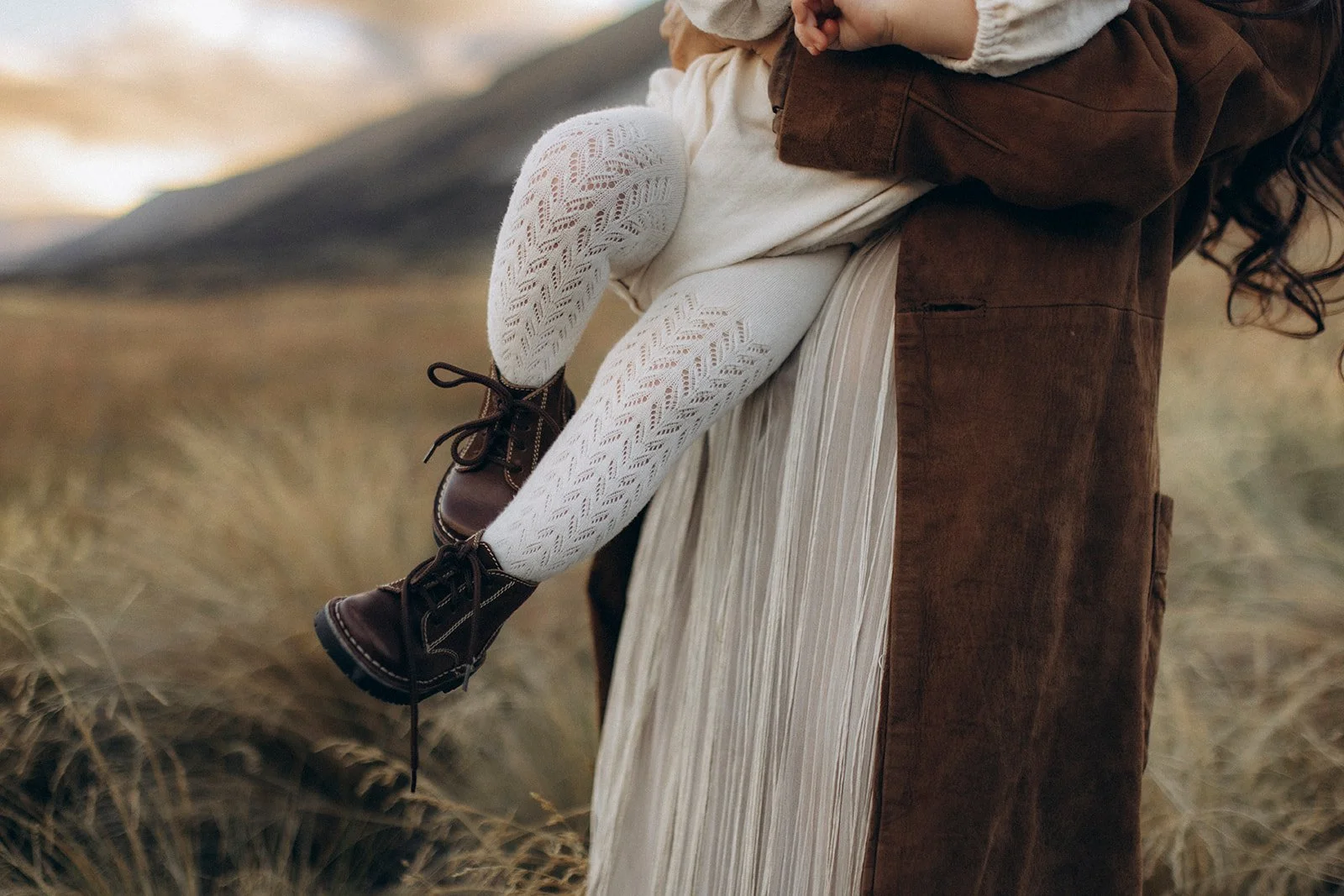 A woman in a brown suede jacket and a cream pleated skirt holds a child in white lace tights and dark brown leather boots, standing in a field of tall grass.