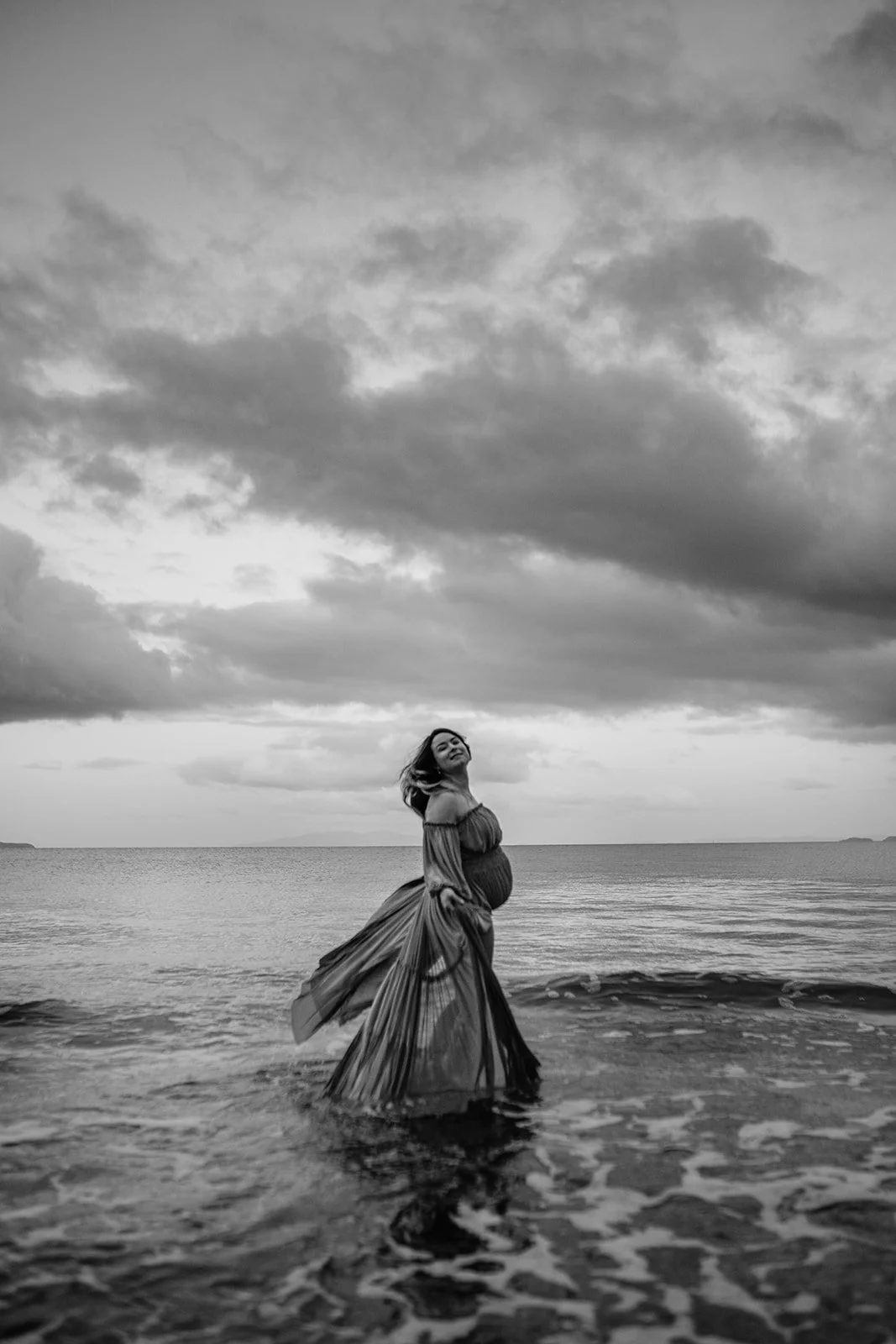 A woman standing in the ocean, wearing a long flowing dress, with a cloudy sky above.