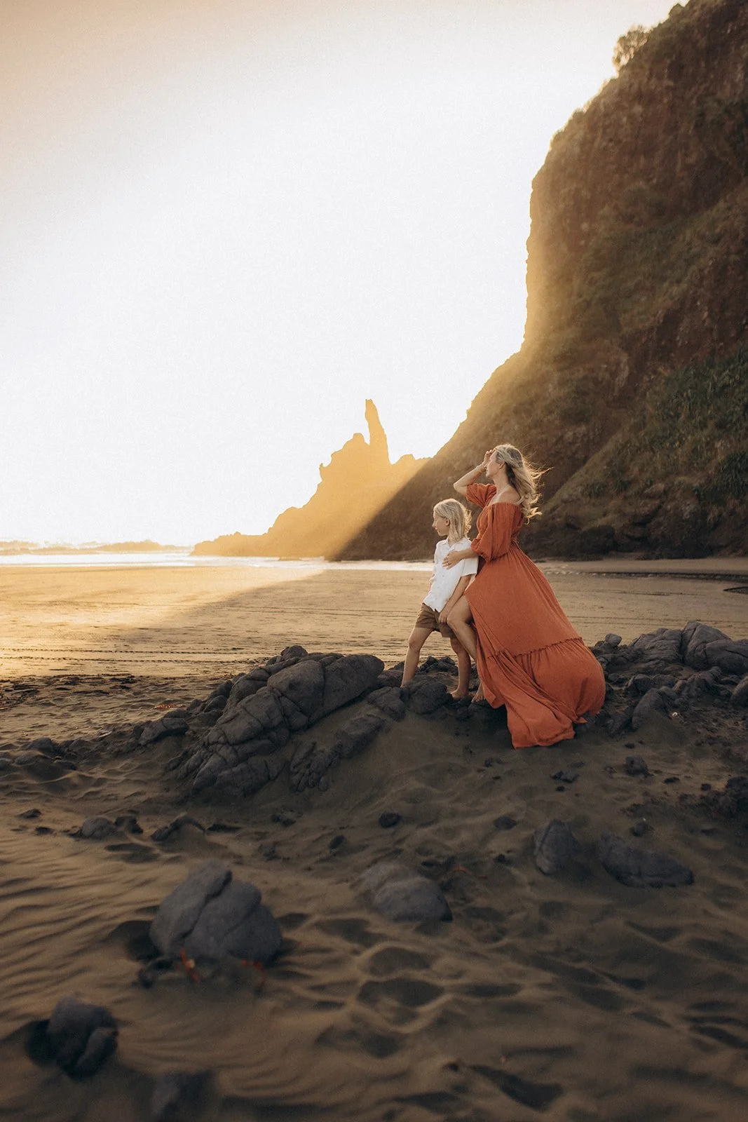 A woman and a young girl sitting on rocks on a beach with cliffs in the background, during sunset.