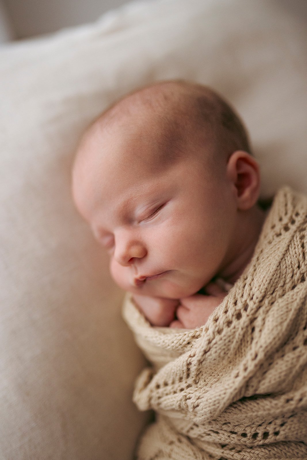 Close-up photo of a sleeping baby with a beige knitted blanket around them, resting on a light-colored pillow.