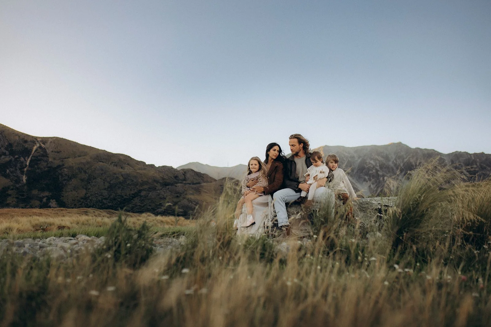 A family of five sitting on a large rock in a field of tall grass with mountains in the background, under a clear sky.