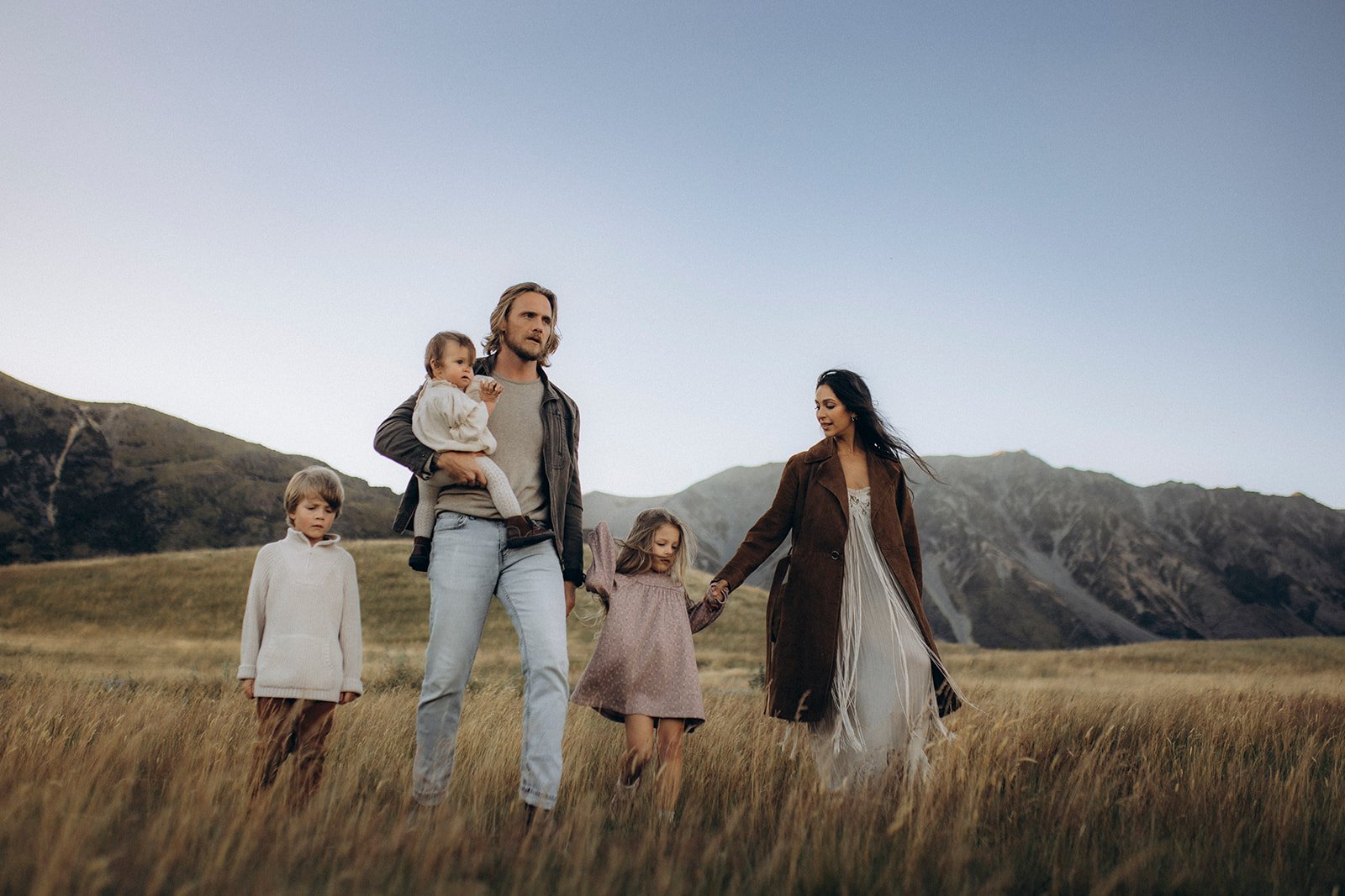 A family of five walking through a grassy field with mountains in the background. The father is holding a young girl, while the mother holds hands with an older girl. A boy walks nearby.