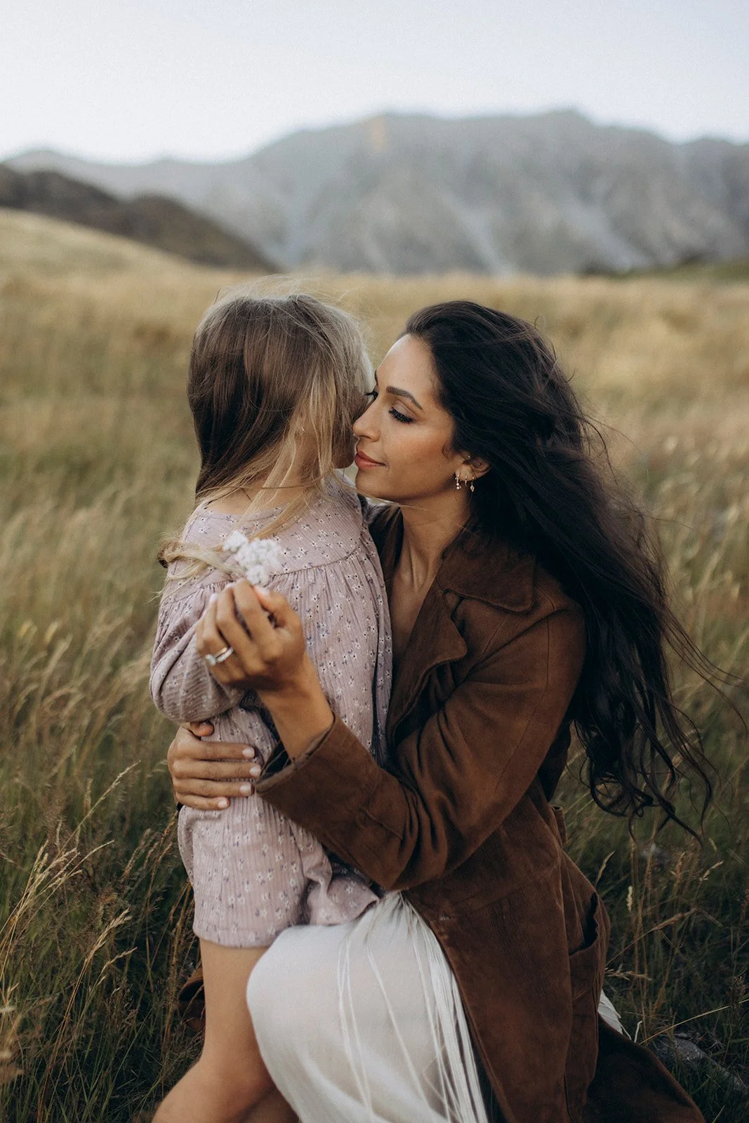 A woman with long dark hair and earrings embraces a young girl with blonde hair in a field with mountains in the background.