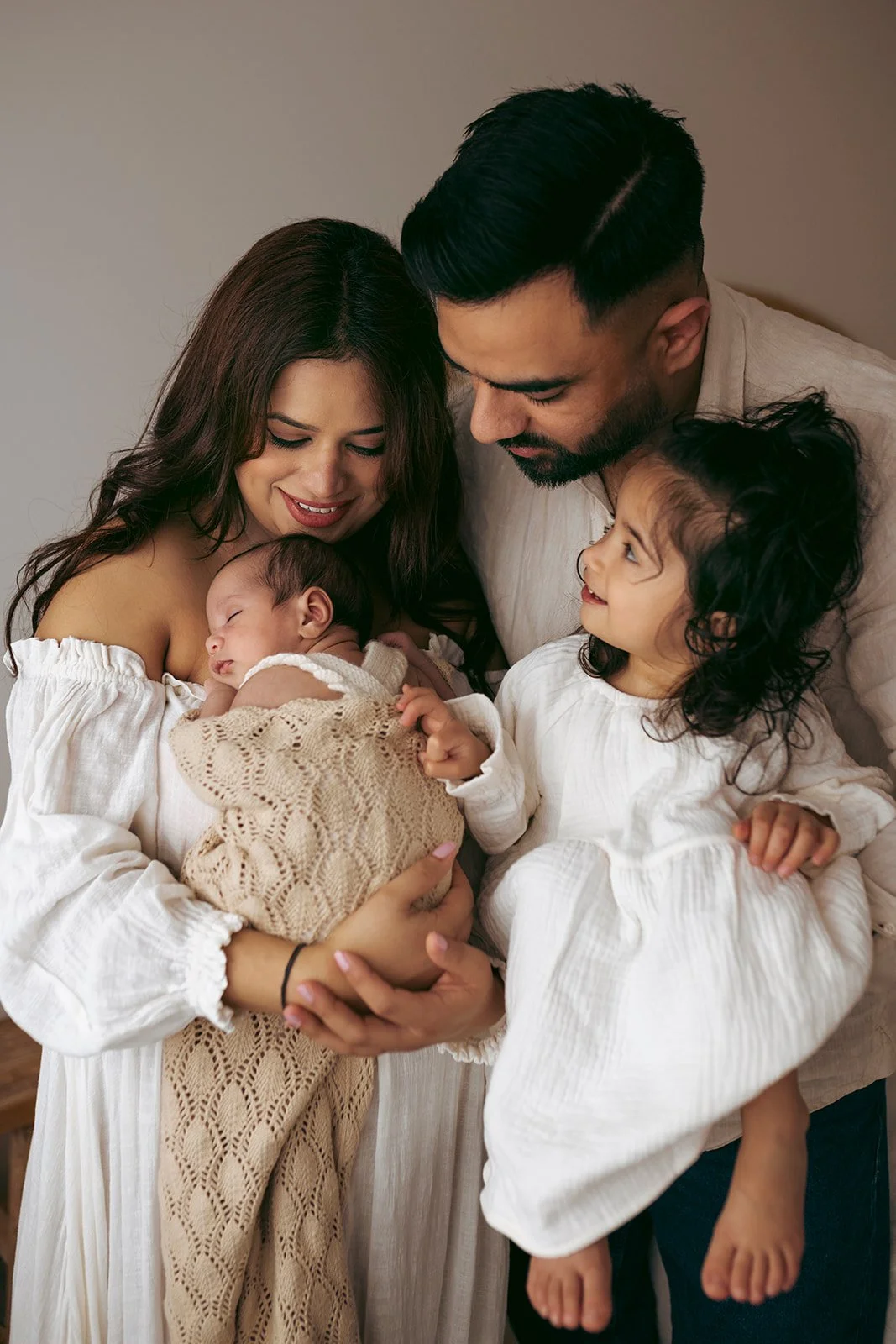 A family of four with a baby, mother, father, and young girl, all dressed in white, sharing a tender moment indoors.