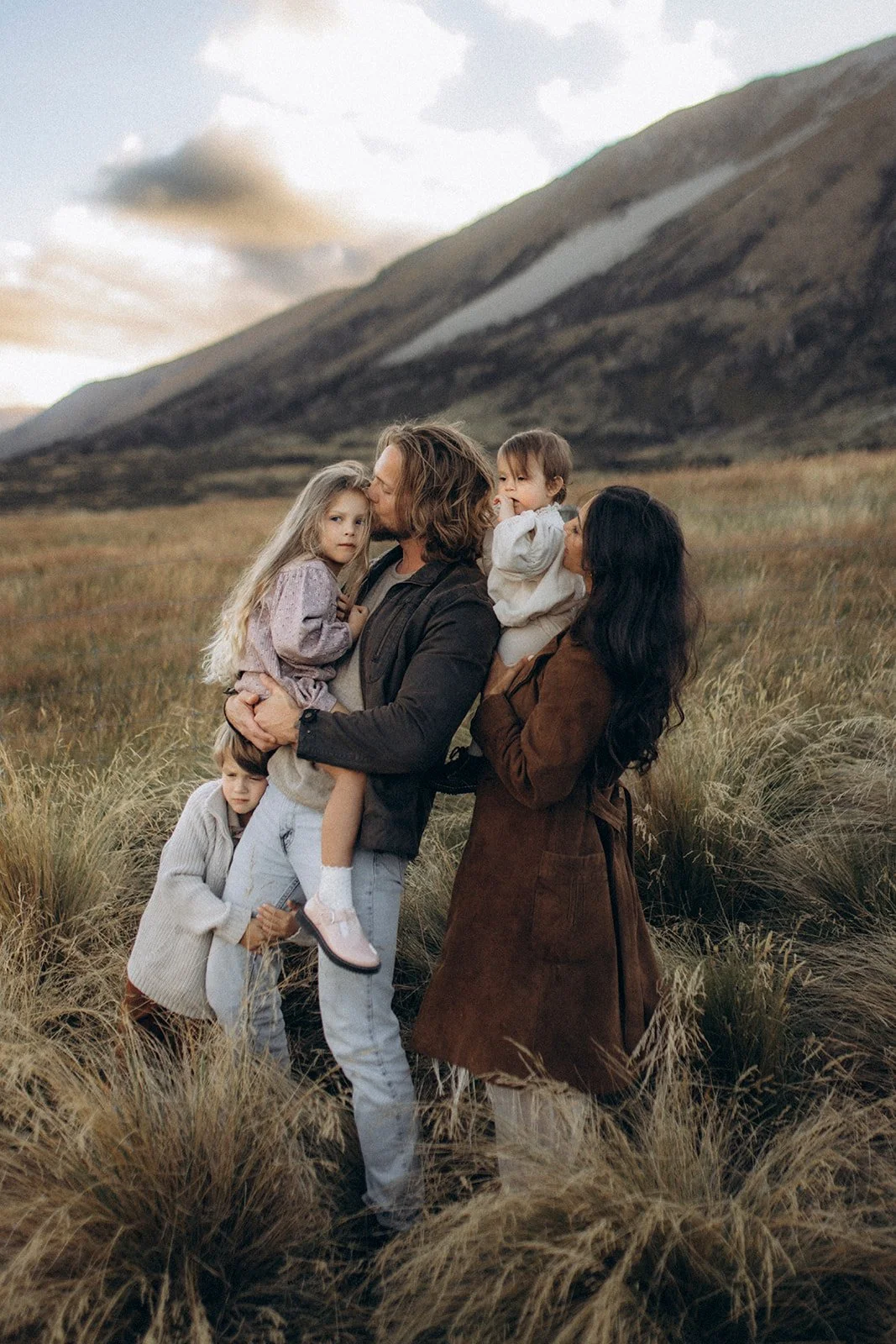 A woman and a man holding two young children in a grassy field with mountains in the background during daylight.