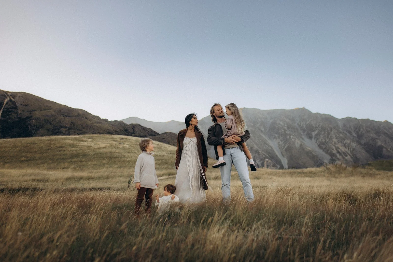 A family standing in a grassy field with mountains in the background. The family includes a man holding a girl, a woman, and two young children, one boy and one girl.