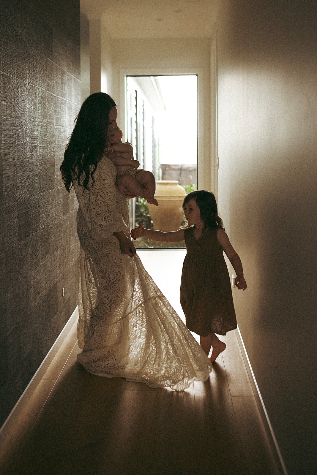 A woman holding a baby in a hallway, a young girl reaching towards the woman, with a bright doorway in the background.