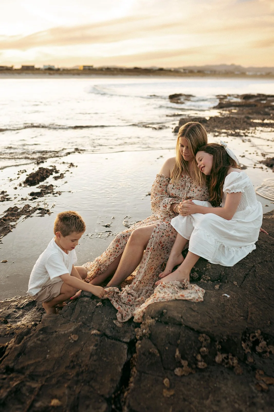 Two young women sitting on rocks at the beach during sunset, with a young boy playing near their feet, and ocean waves in the background.