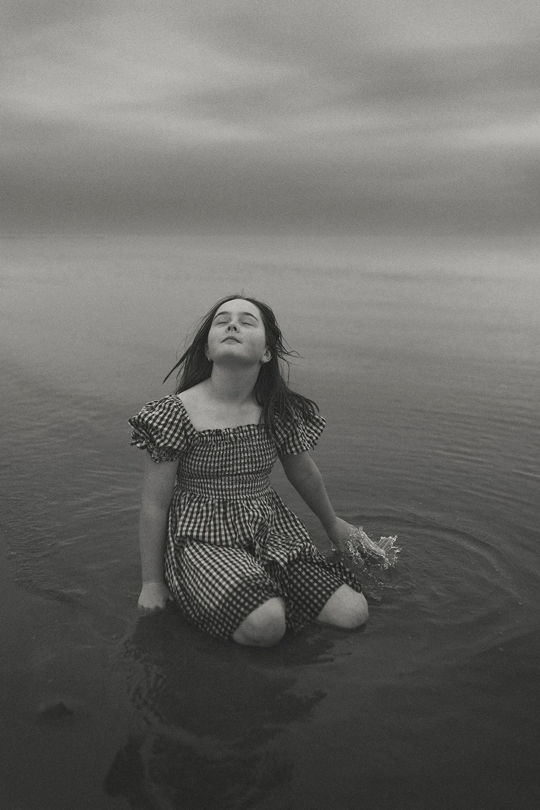 Black and white photo of a young woman with long hair, sitting knee-deep in water with her eyes closed and face tilted upward, wearing a checkered dress.