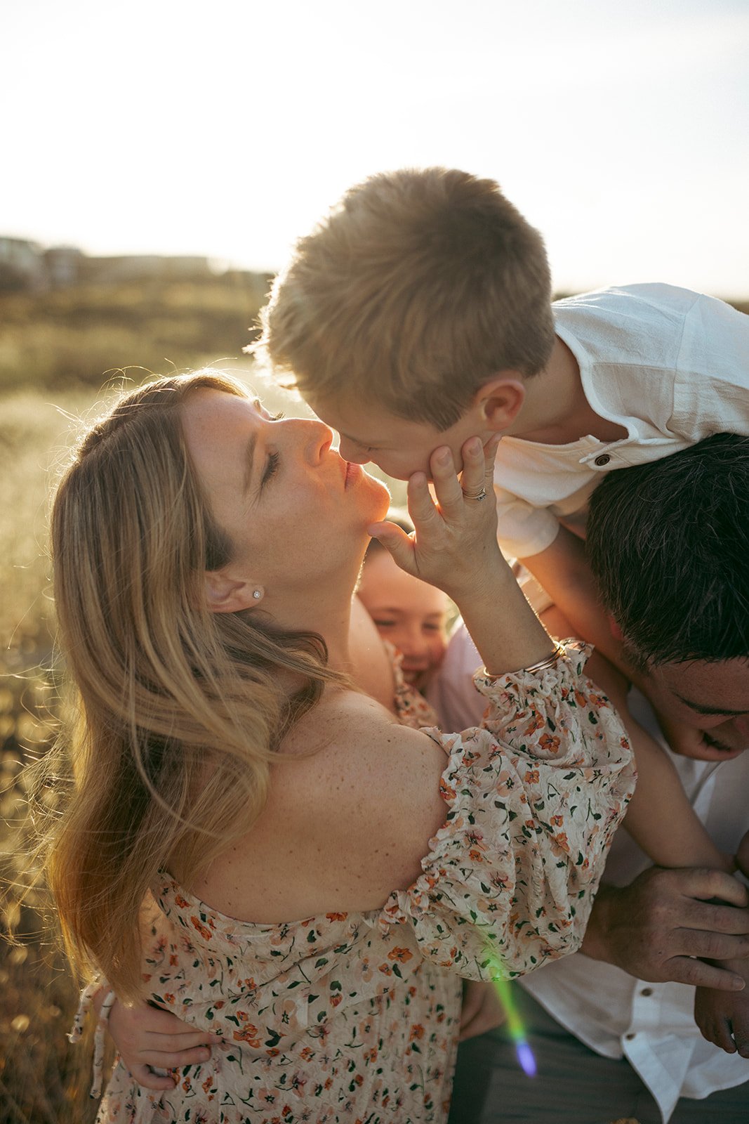 Family outdoors at sunset, with a woman and a man holding their children, sharing an intimate moment, close-up shot.