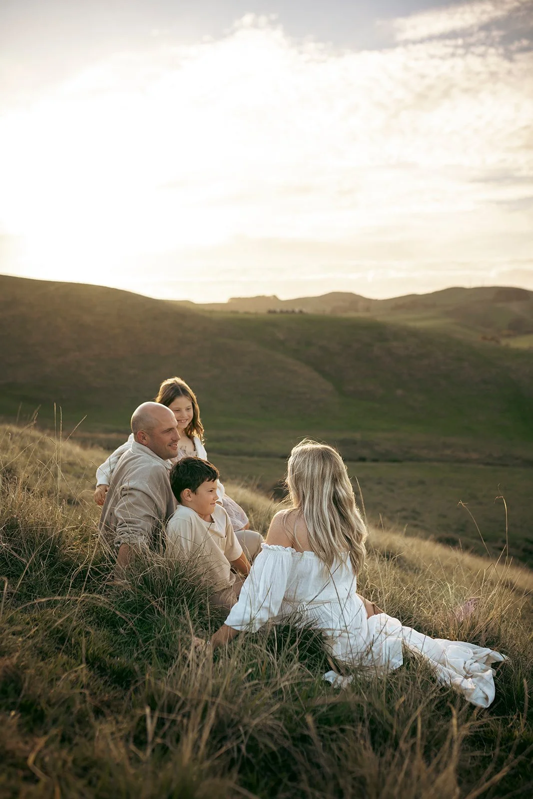 Family of four sitting on a grassy hillside, enjoying a sunset view of rolling hills.