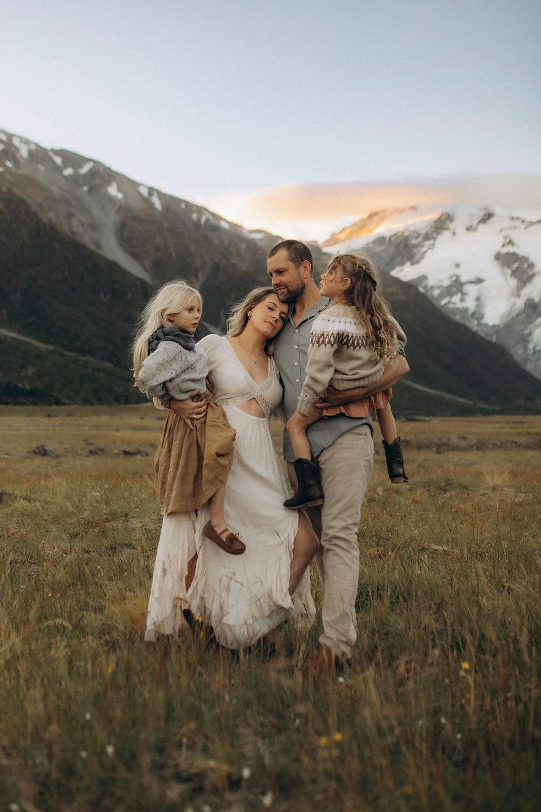 A family of four, including two young girls, standing together in a grassy field with mountains in the background during sunset.