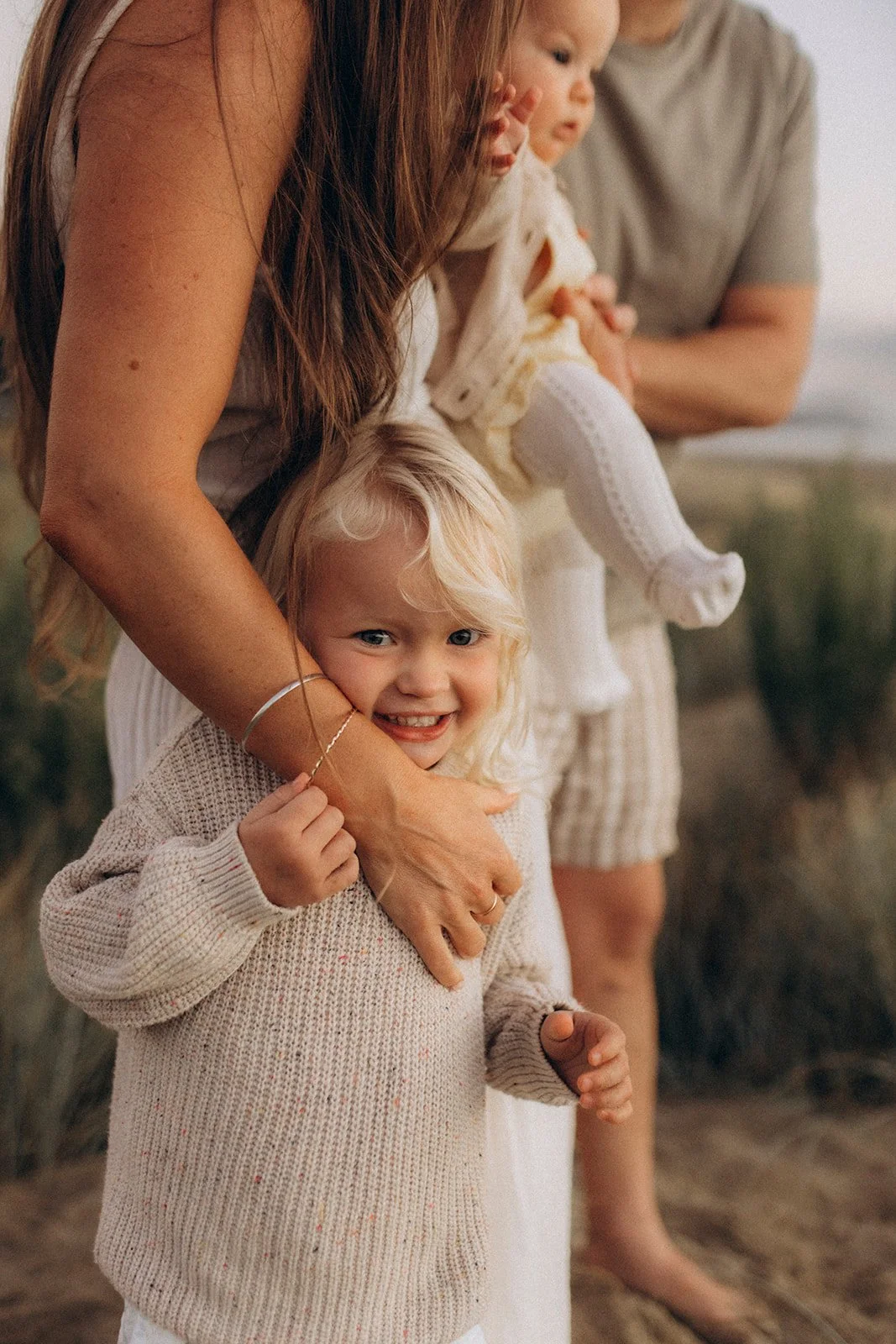 A smiling young girl with blonde hair and a beige sweater hugging an adult woman's waist, standing outdoors near a beach or coastal area, with two other children and an adult in the background.