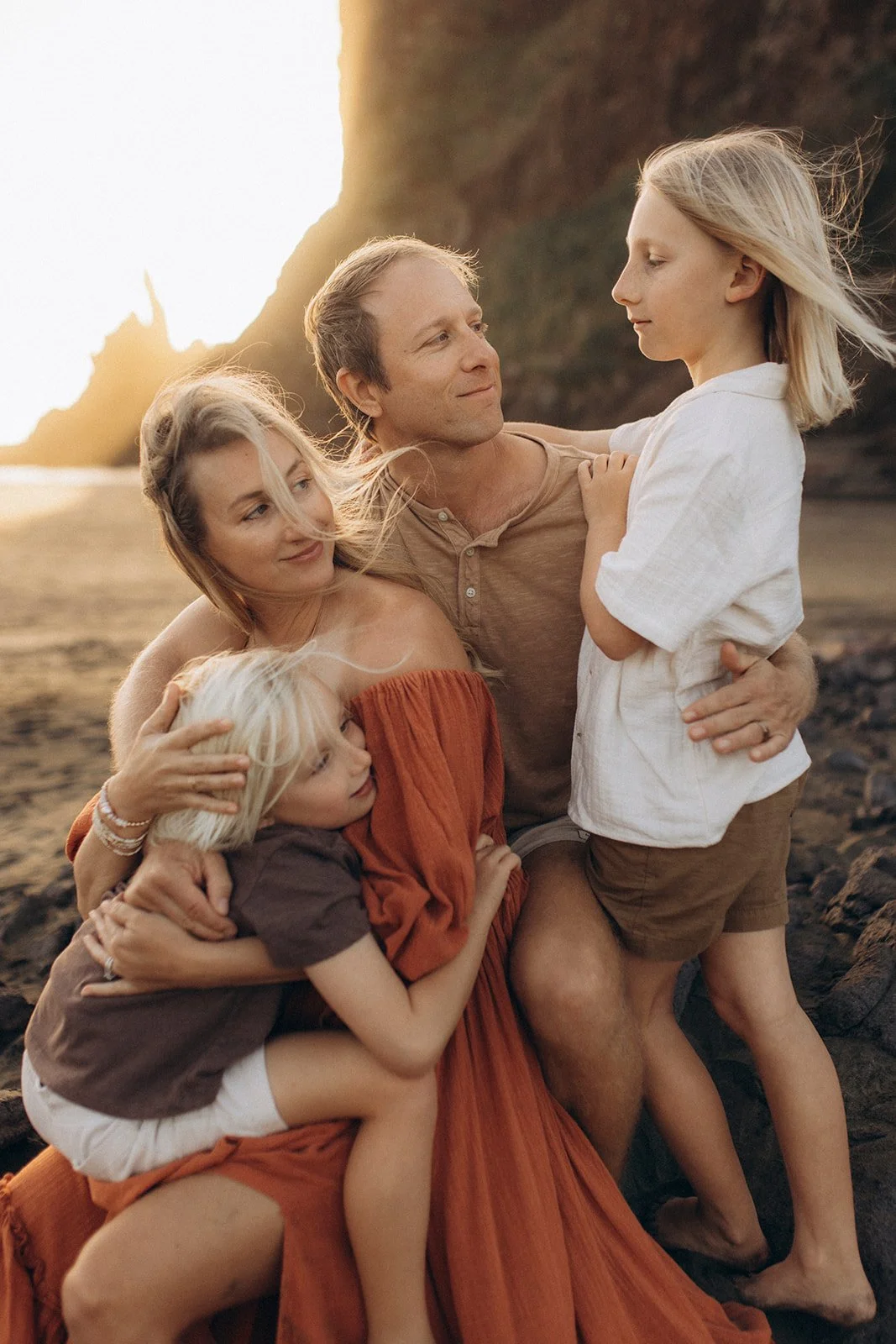 A family of five on the beach at sunset: a father, mother, two young daughters, and a young son, embracing near large rocks with a cliff in the background.