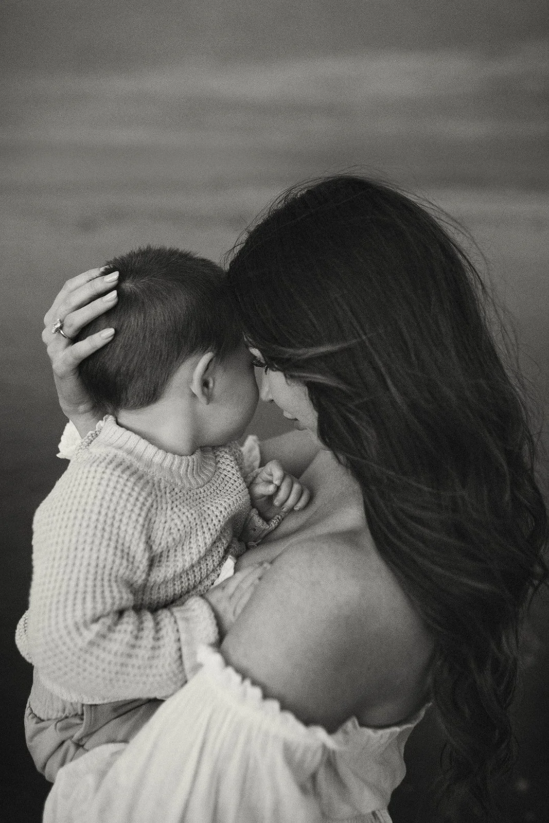 A woman with long dark hair gently holding a young child with a short haircut close to her face, both with their eyes closed, in a tender embrace.