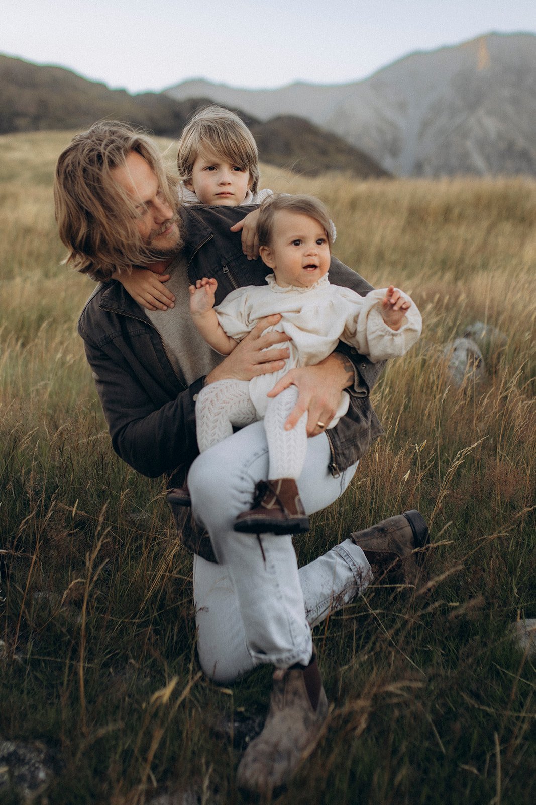 A man with long, wavy hair holding two young children on a grassy field with mountains in the background.