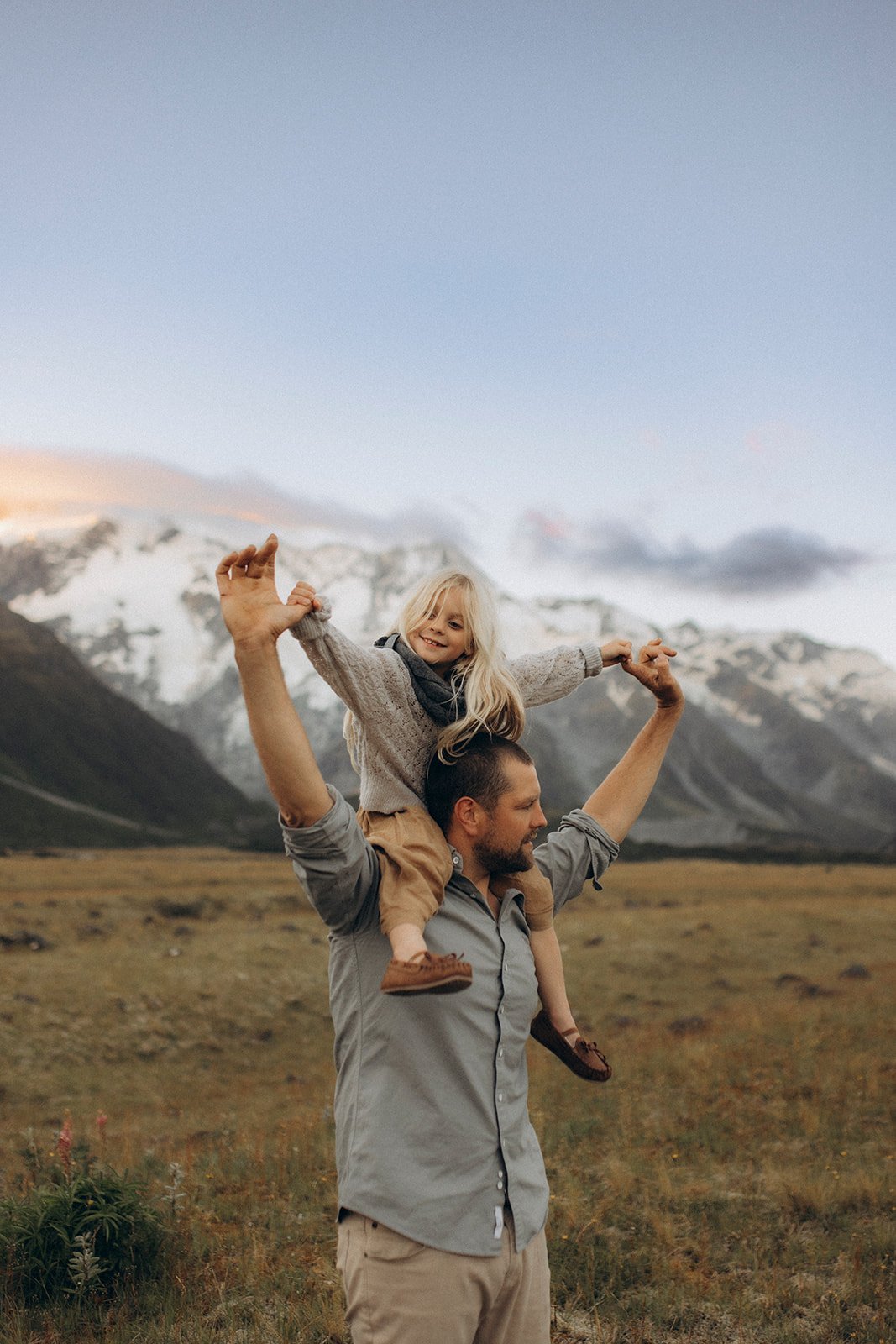 A man carrying a girl on his shoulders in a mountain landscape with snow-capped peaks and a cloudy sky.