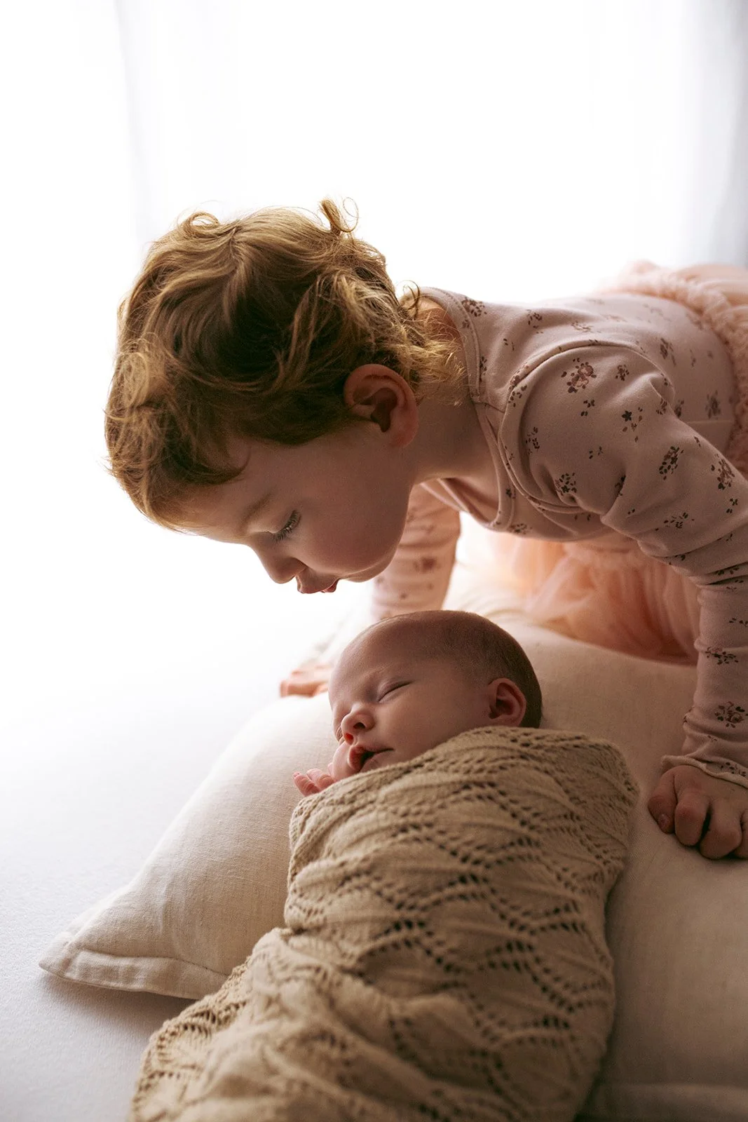 A young girl leaning over a baby who is sleeping on a pillow, holding a blanket, with soft natural light in the background.