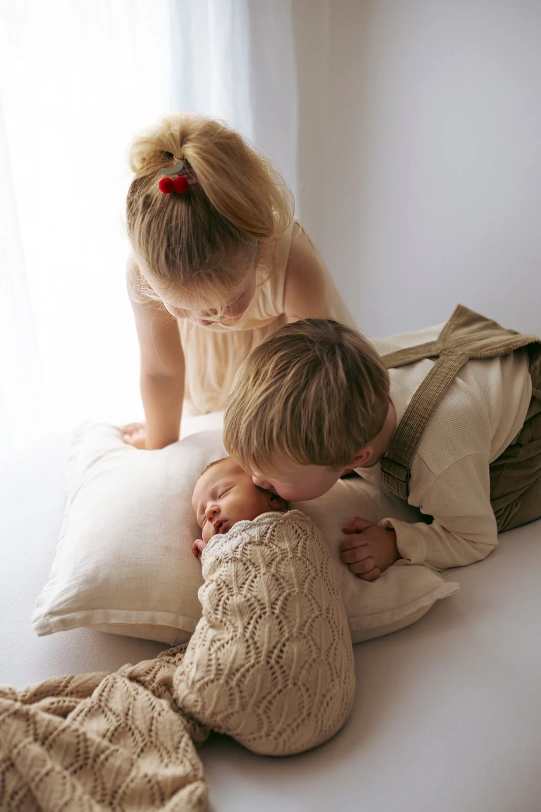 A young girl and an older boy with light hair are leaning over a newborn baby who is sleeping on a pillow on a bed, in a softly lit room.