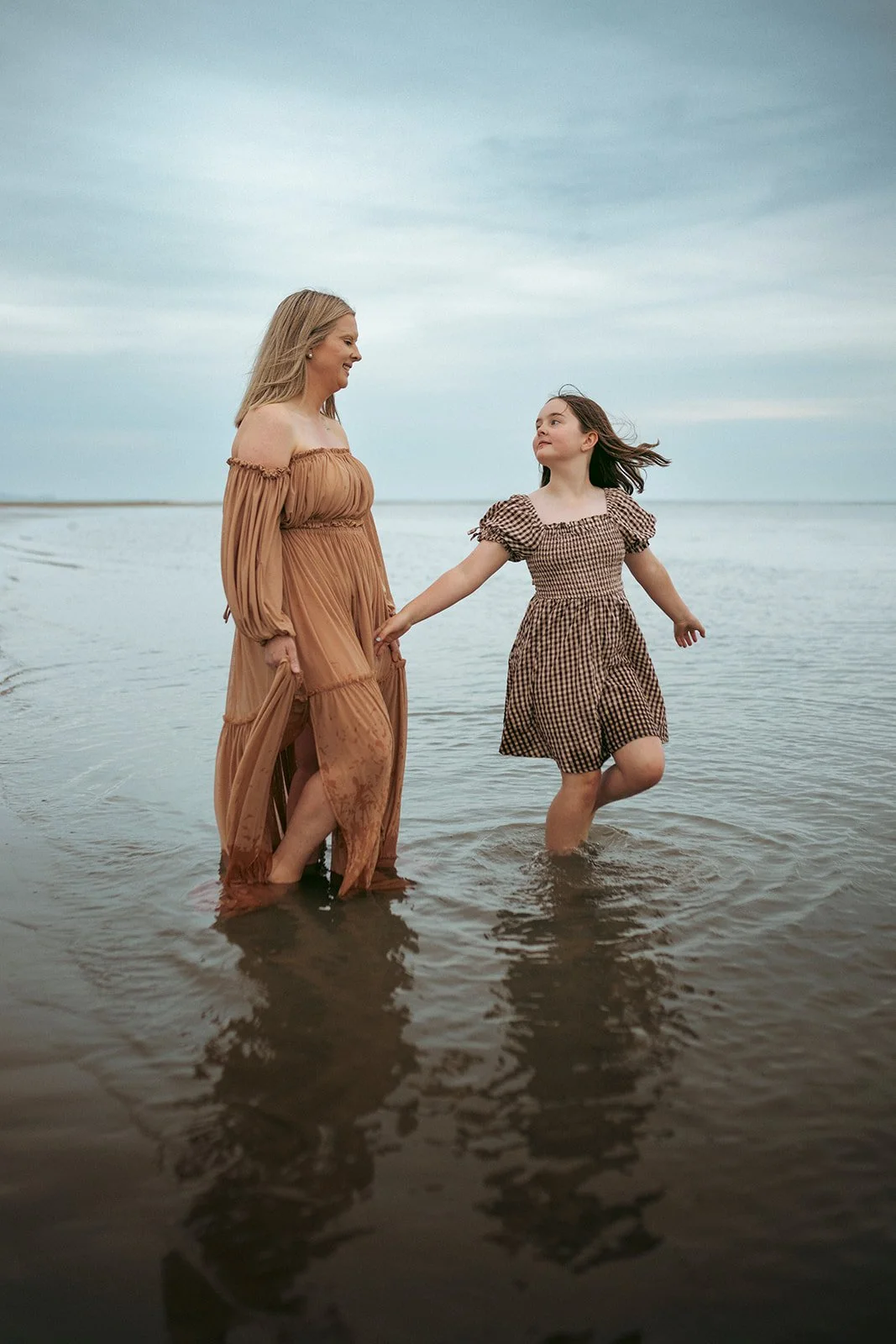 A woman and a girl holding hands while standing in shallow water at the beach, both smiling and looking at each other.