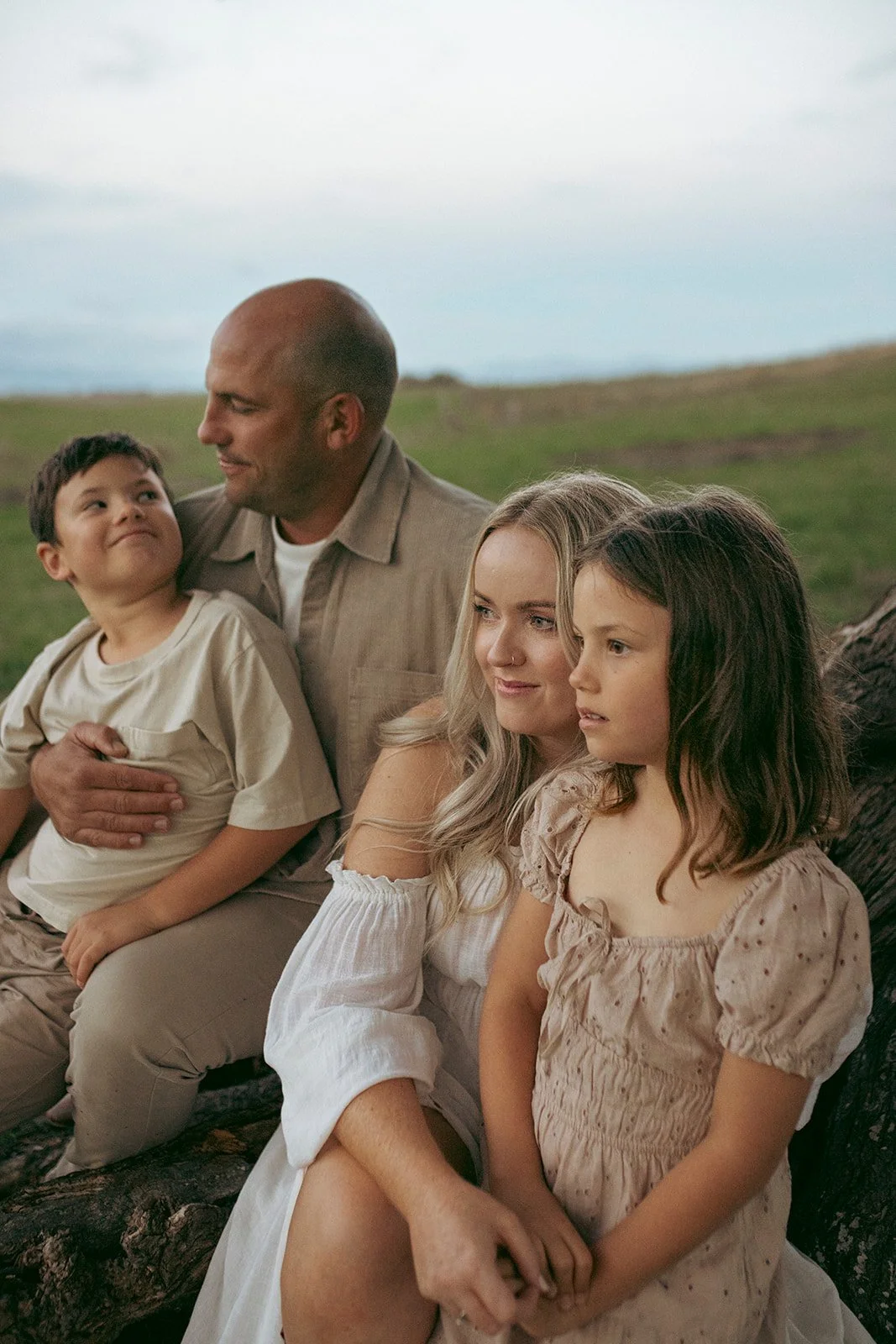 A family of four sitting outdoors on a log in a grassy field, with a cloudy sky in the background. The father, an adult male, is holding a young boy, while two young girls sit beside them.