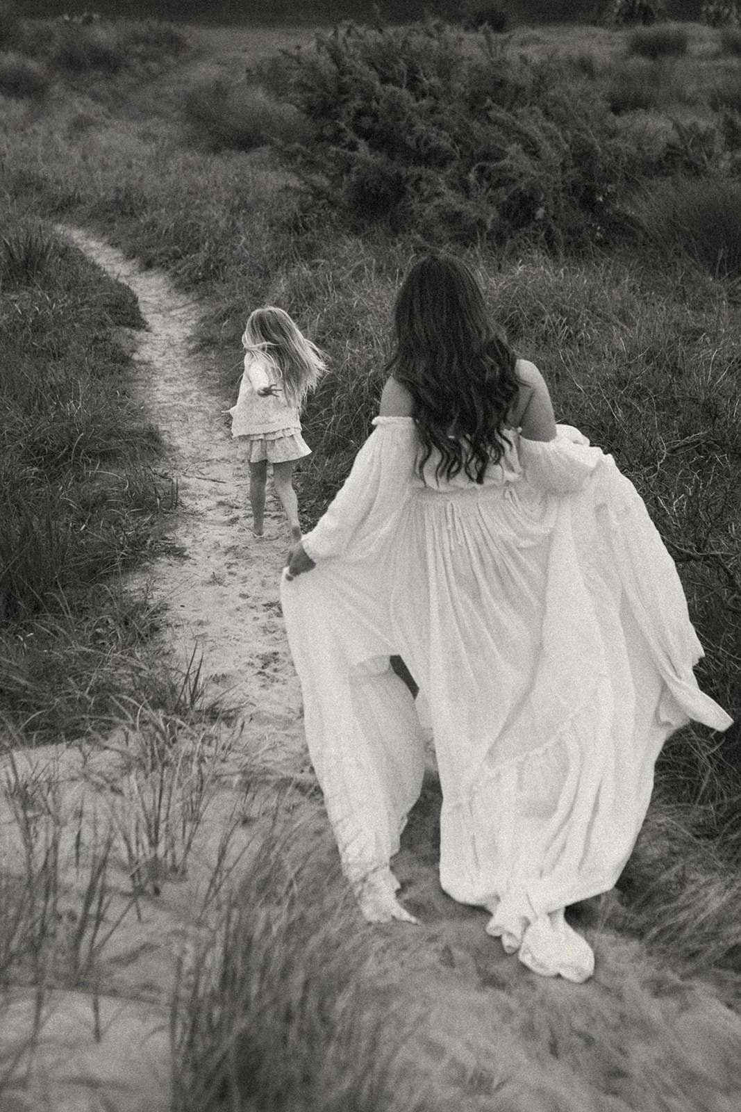 A woman in a flowing white dress walking on a sandy trail through tall grass, with a young girl with long hair in a light dress walking ahead on the same trail, in a natural outdoor setting.