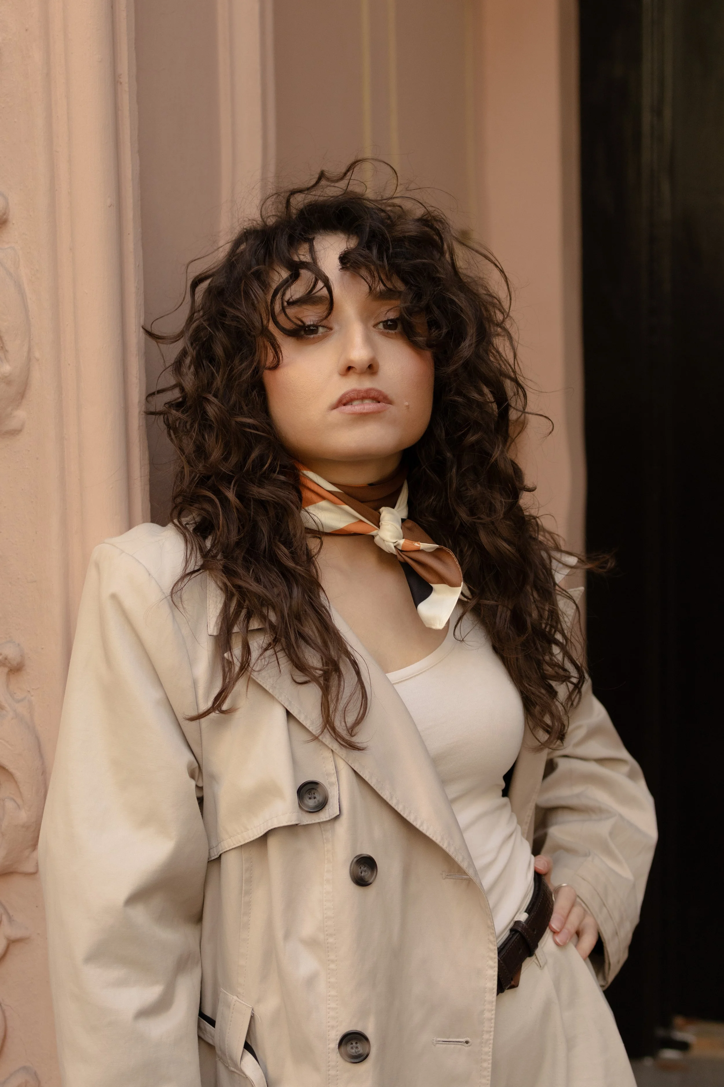 A young woman with curly brown hair wearing a beige trench coat, a white top, and a light brown and white scarf around her neck, standing against a wall with her hand on her hip, looking at the camera.