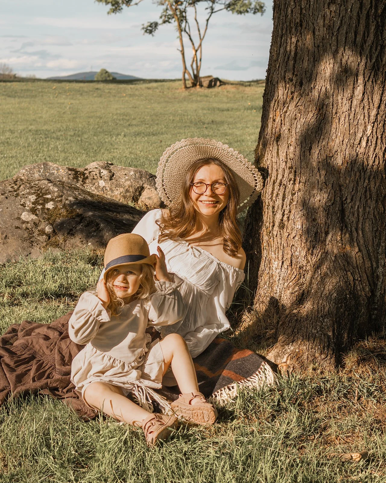 A woman and a young girl sitting on grass under a large tree, both wearing sun hats and smiling, enjoying a sunny day outdoors.