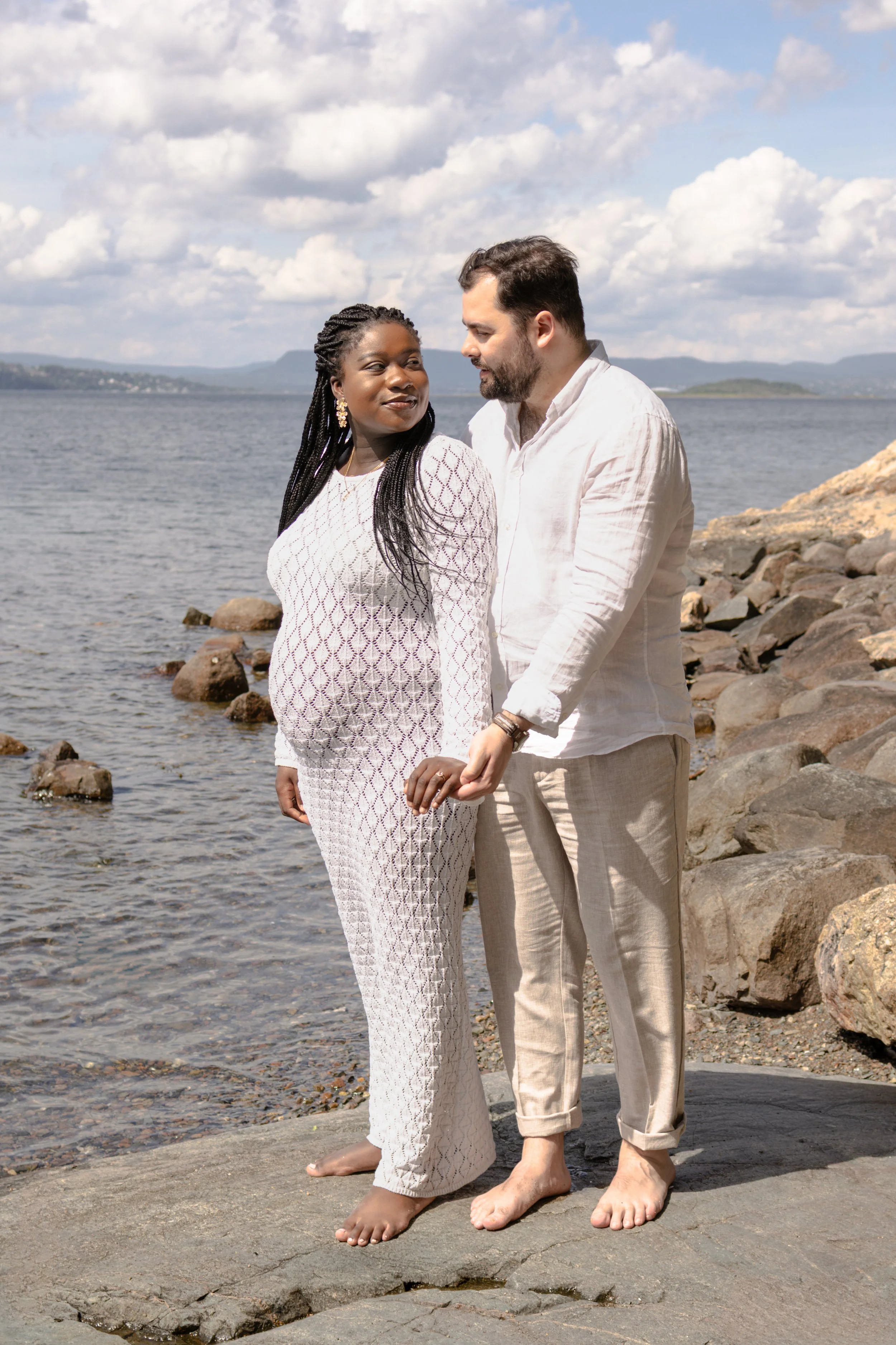 A couple standing on a rocky shoreline near a body of water with mountains in the background. They are holding hands, barefoot, dressed in light-colored, casual clothing. The woman is wearing a patterned dress with long sleeves and jewelry, while the man is wearing a linen shirt and pants. The sky is partly cloudy.