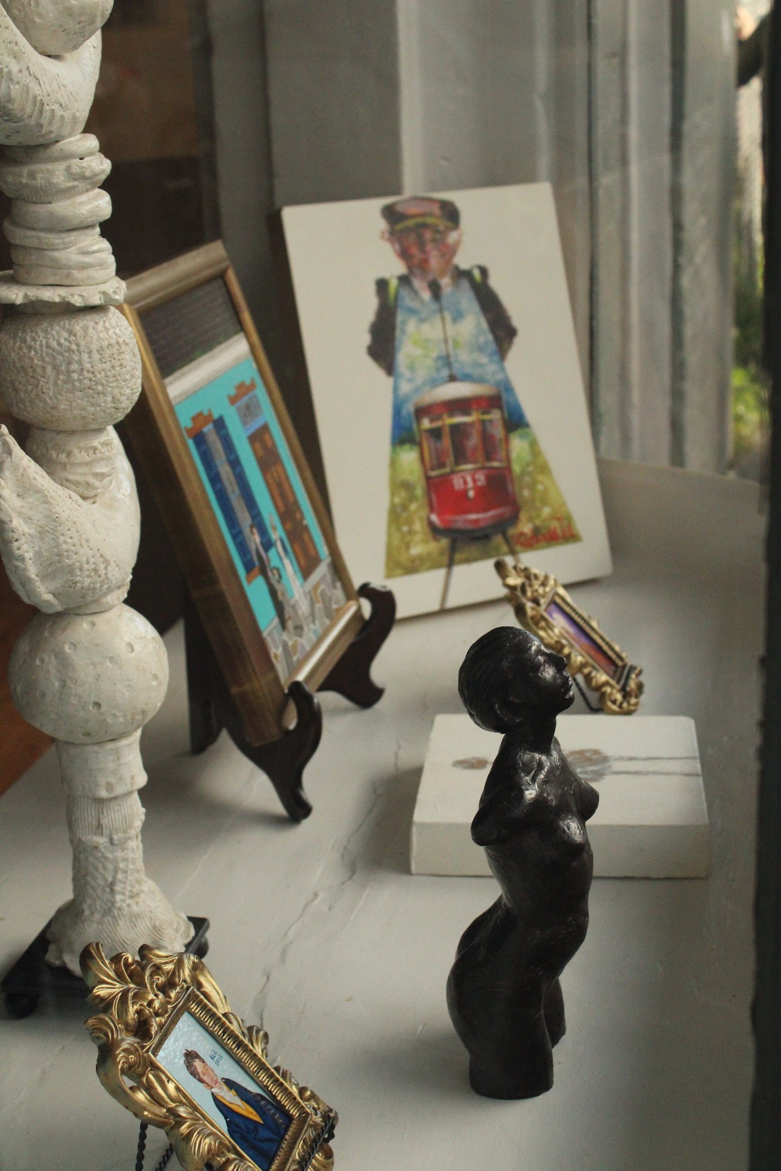 A shelf displaying framed photographs, sculptures, and decorative items, including a small black bust and a figurine, against a wall with a window.