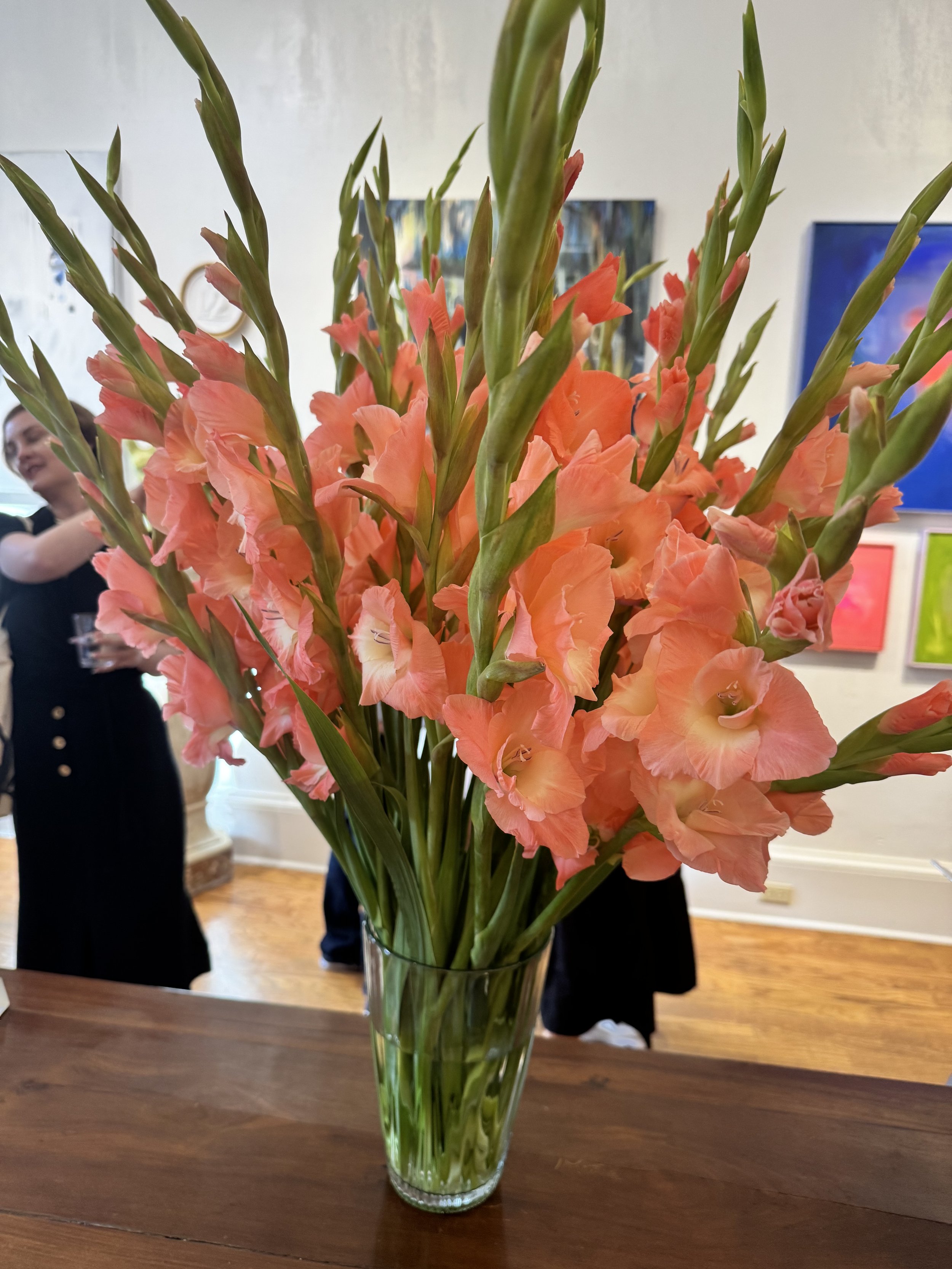 Pink gladiolus flowers in a clear glass vase on a wooden table, with people and artwork visible in the background.