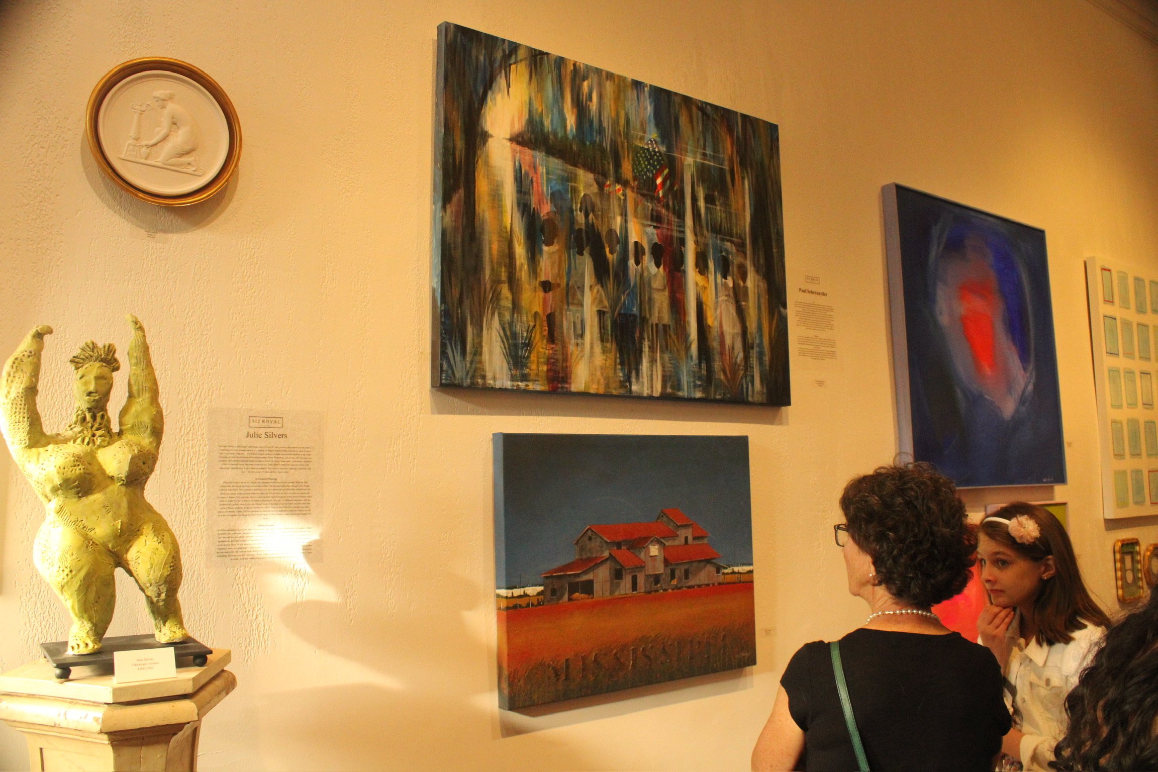 People observing paintings and artwork in an art gallery or museum. The wall displays several colorful paintings, and there is a sculpture of a person on a pedestal on the left.