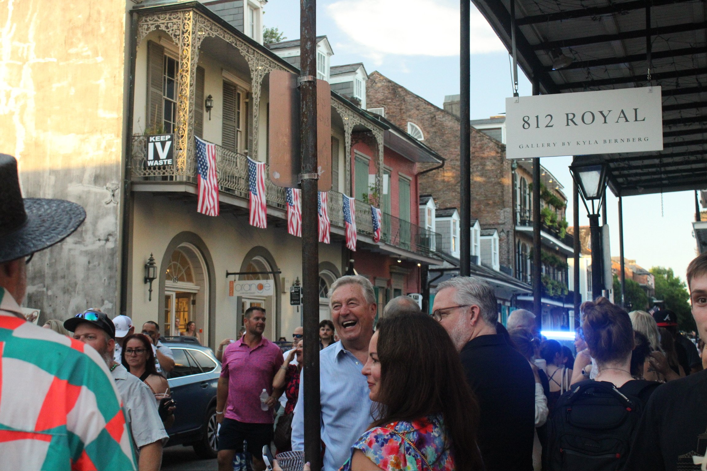 A crowd of people gathered on a city street during a celebration or event, with buildings and American flags in the background. Some individuals are smiling and talking.