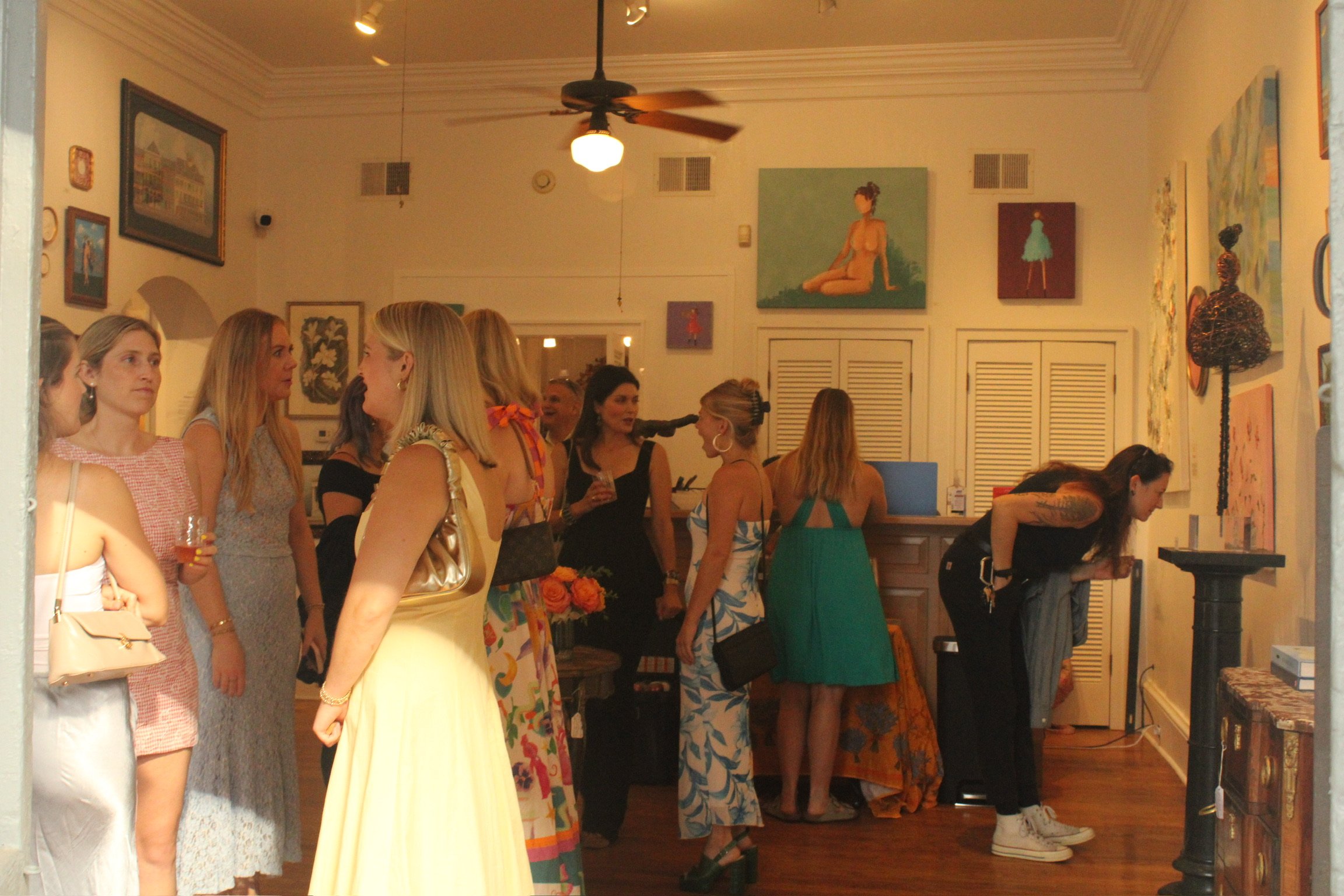 People gathered in an art gallery or studio space, engaging in conversation and viewing artwork. The room features various paintings on the walls, and a ceiling fan is visible.