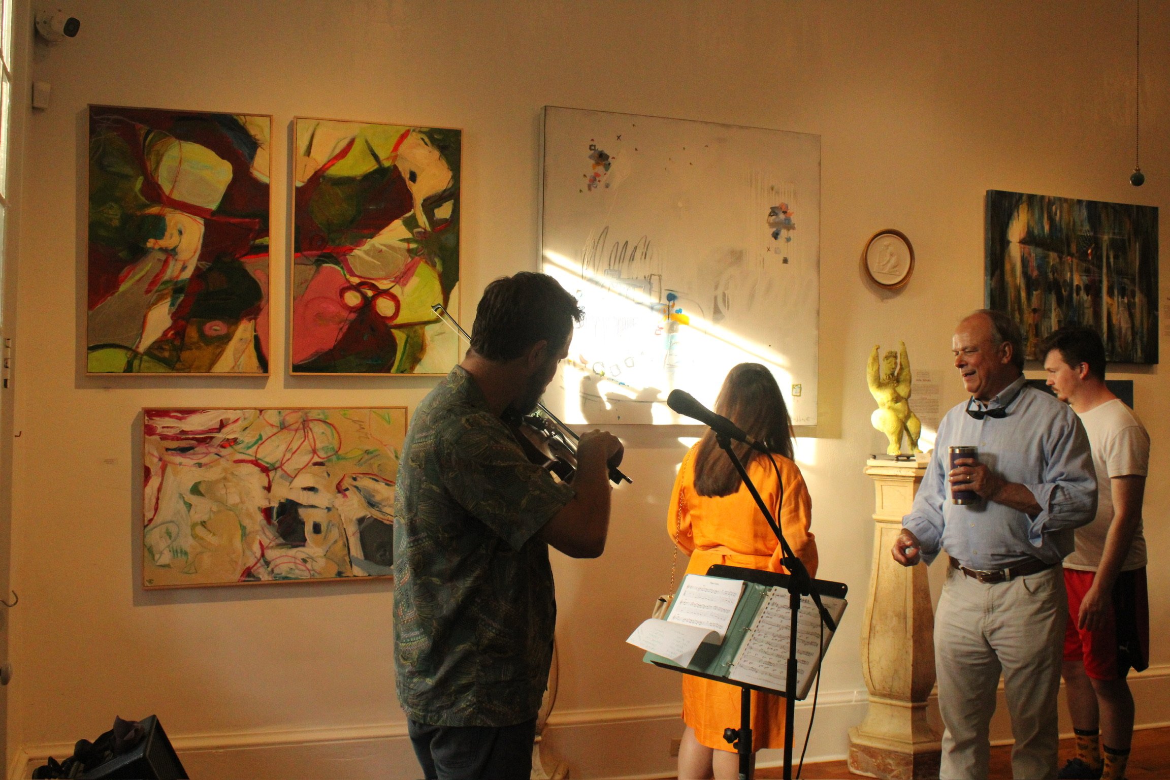 People attending an art gallery event, viewing and discussing abstract paintings on the wall, with some individuals engaging in conversation and music performance.